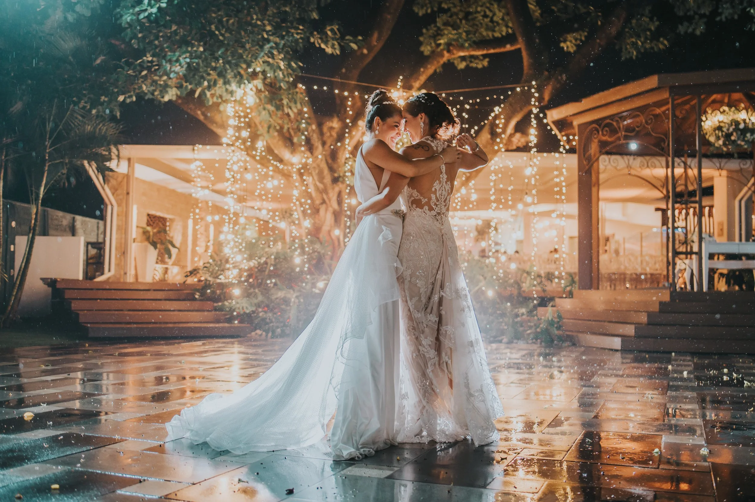 Two women in wedding dresses sharing a dance at an outdoor wedding reception at night, decorated with string lights, in a garden setting.