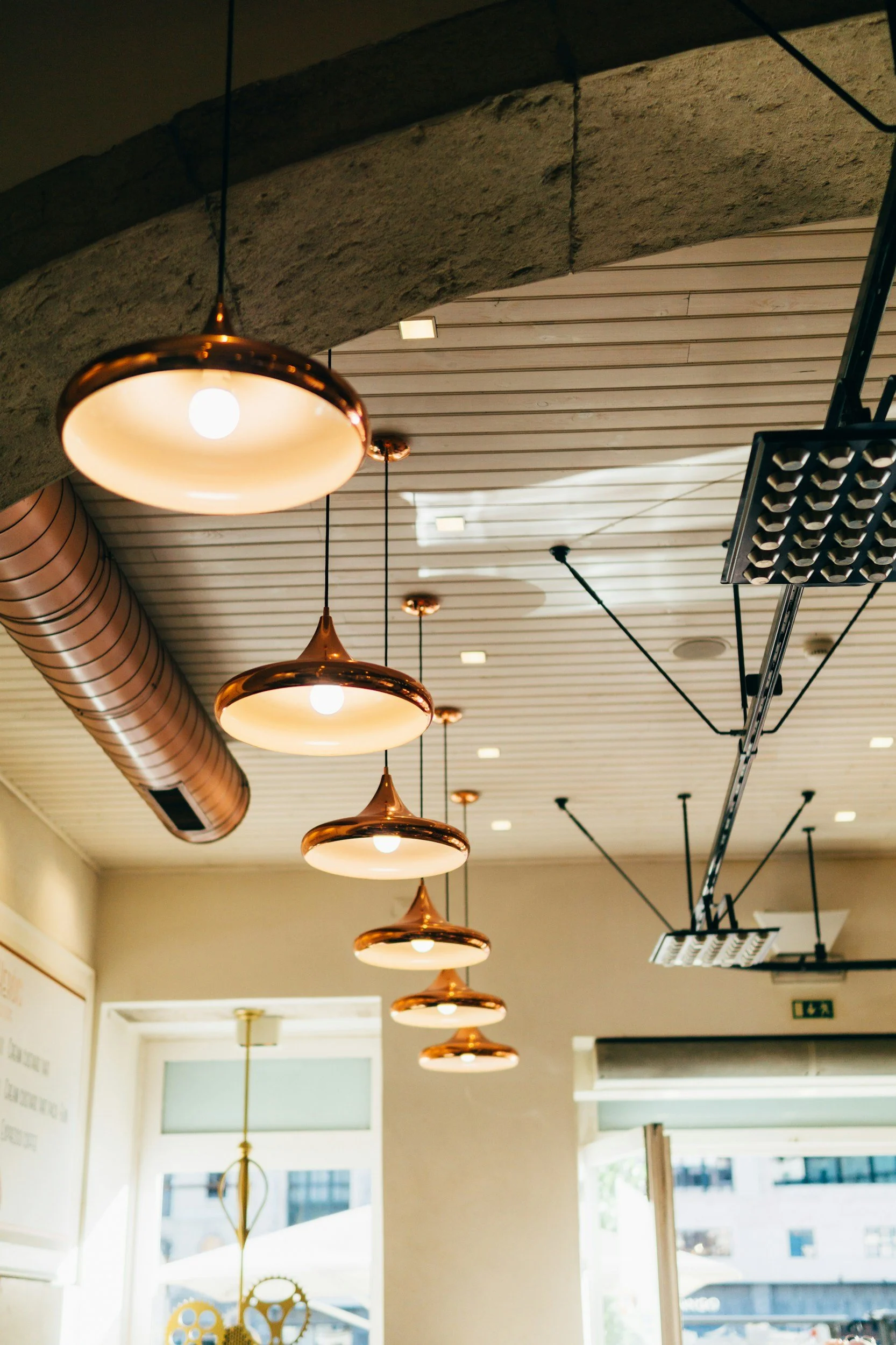 Interior view of a cafe or restaurant with hanging pendant lights featuring copper-colored rims, a building with glass windows outside, and some shelves with gear or clock parts.