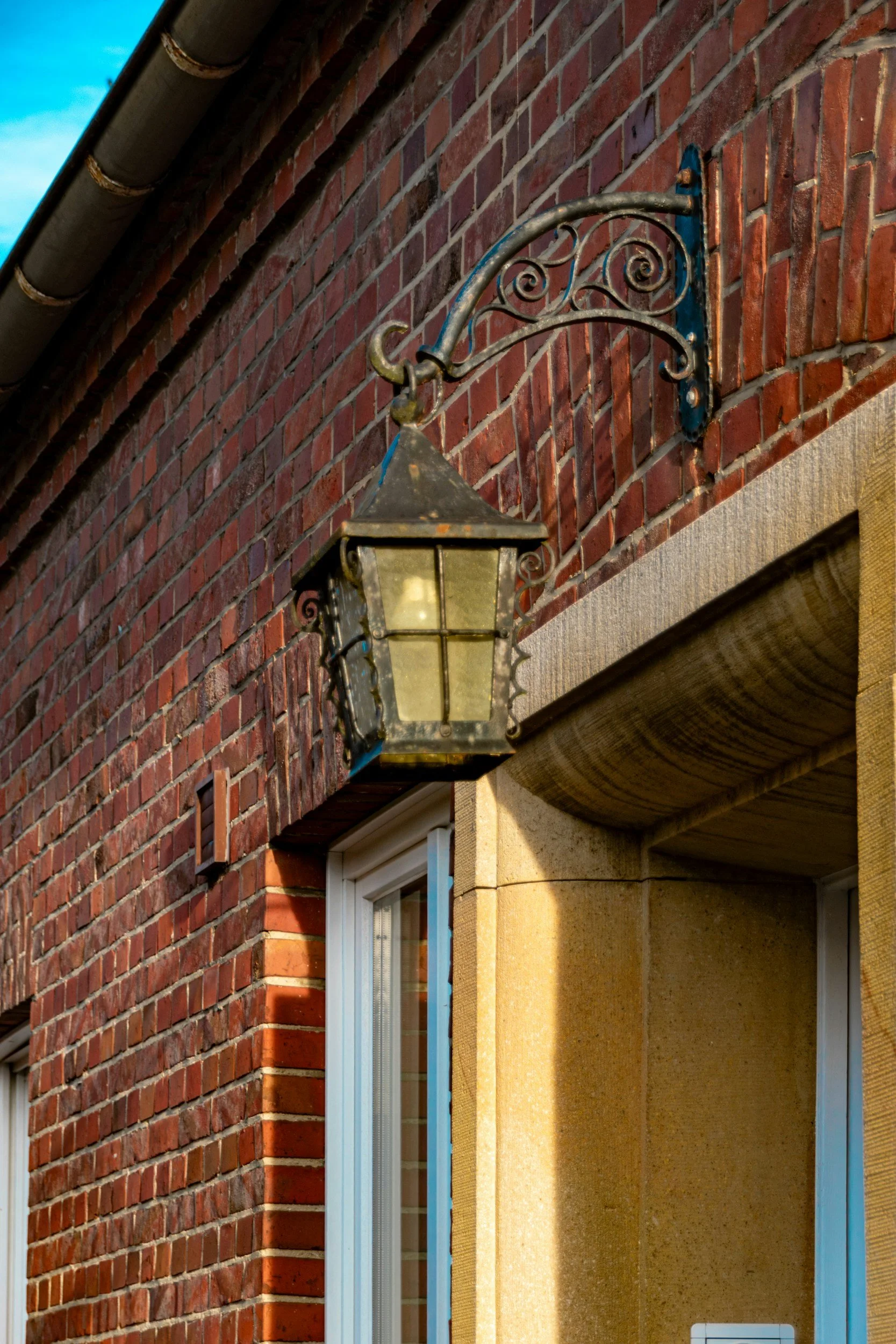A decorative street lamp mounted on a brick wall, with a curved iron arm and ornate details, situated near a window with a beige stone frame.