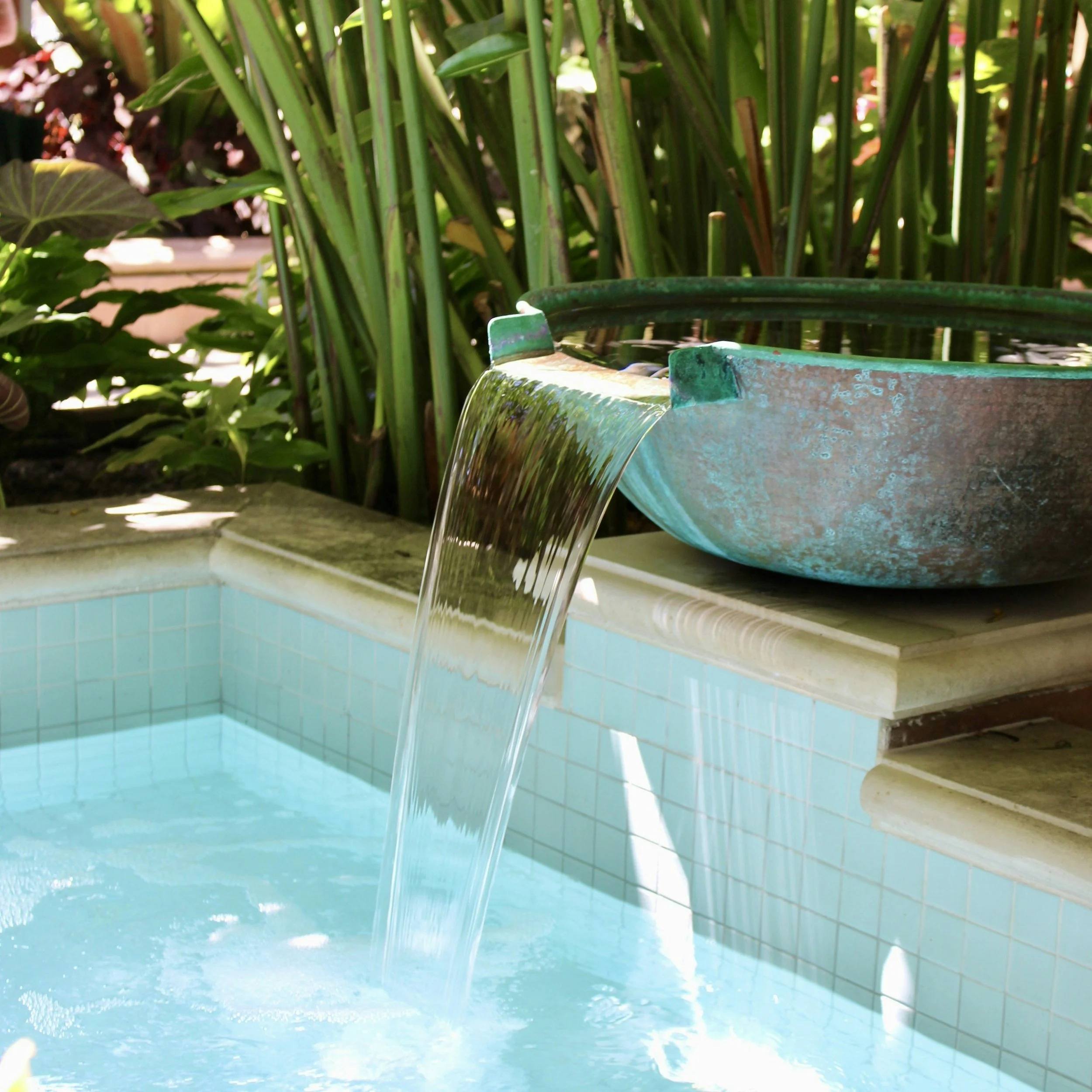 Water cascading from a fountain with lime green tiled walls and lush green plants in the background.