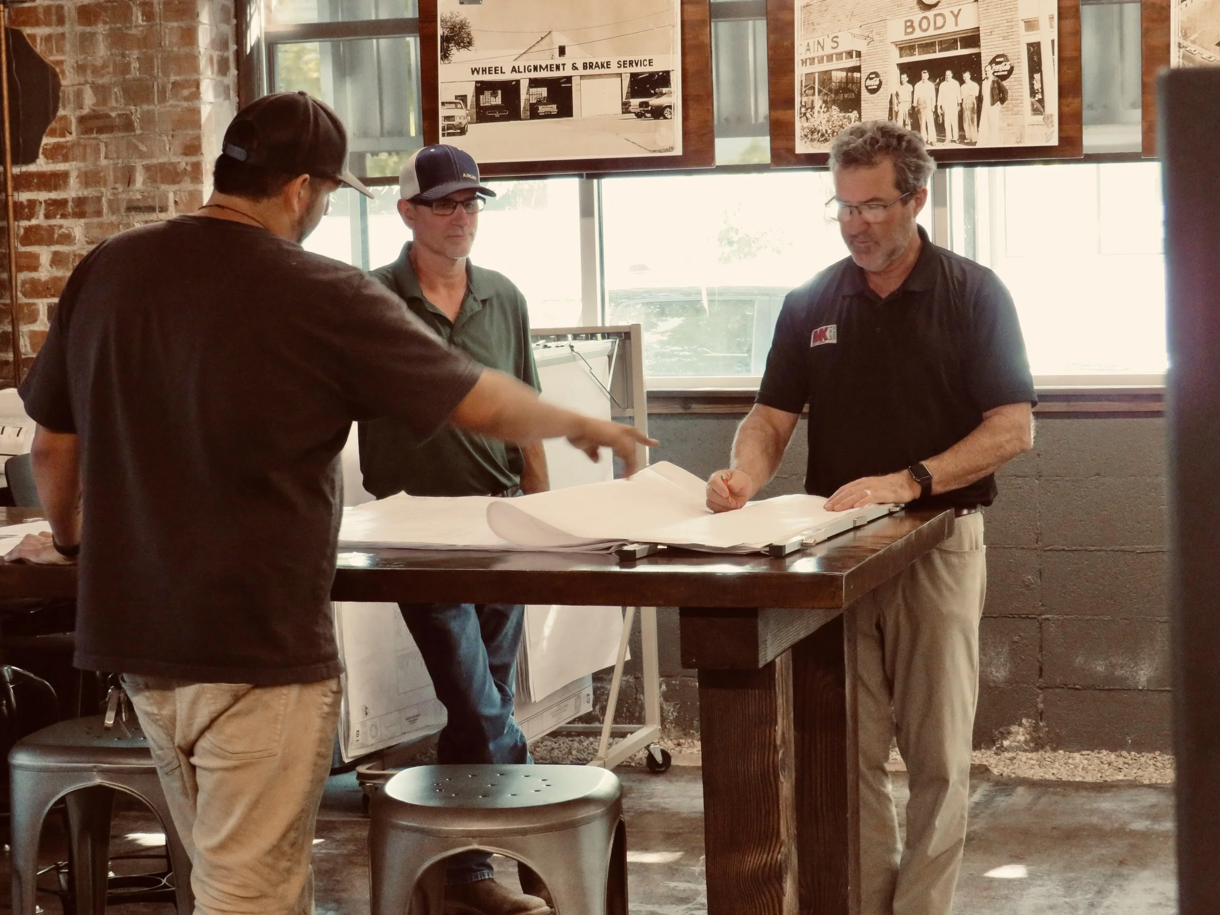 Three men are gathered around a large set of blueprints at a wooden table in a rustic workshop. One man is pointing at the blueprints, while the other two are observing. The workshop has brick and cinder block walls, large windows, and vintage automotive posters.