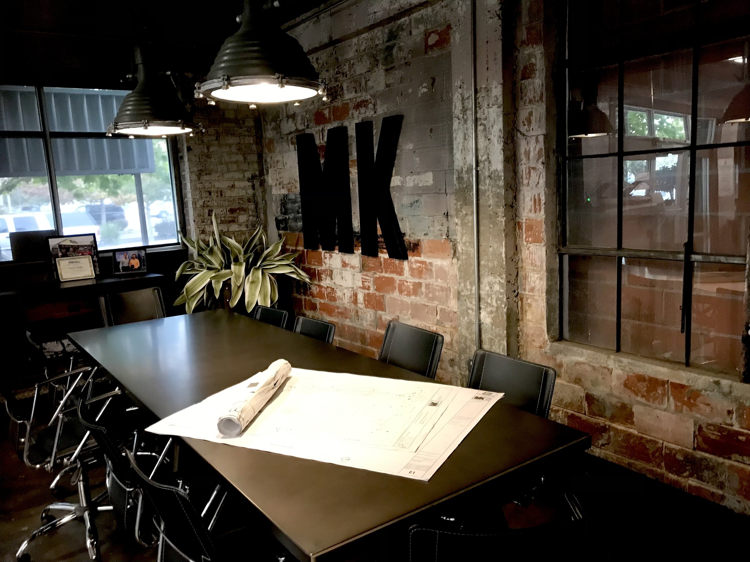 Interior of a meeting room with a long black table, black chairs, exposed brick walls, a window, and large black letters on the wall spelling 'NK'.
