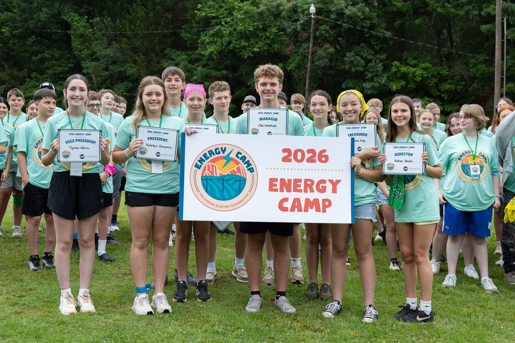 Smile of group of children and teenagers at energy camp holding signs indicating leadership roles, standing on grass with trees in the background.