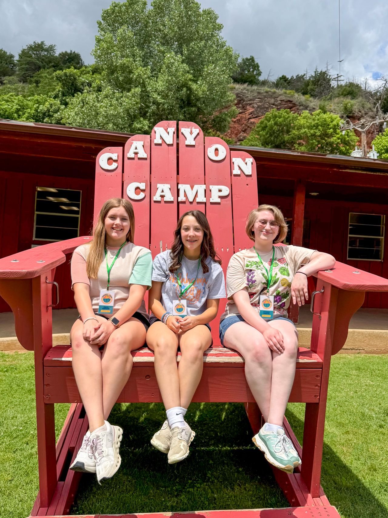 Three young women sitting on a large red bench shaped like a vintage truck at Camp Koronnong, with a sign reading "ANYON CAMP" behind them, surrounded by green trees and a cloudy sky.
