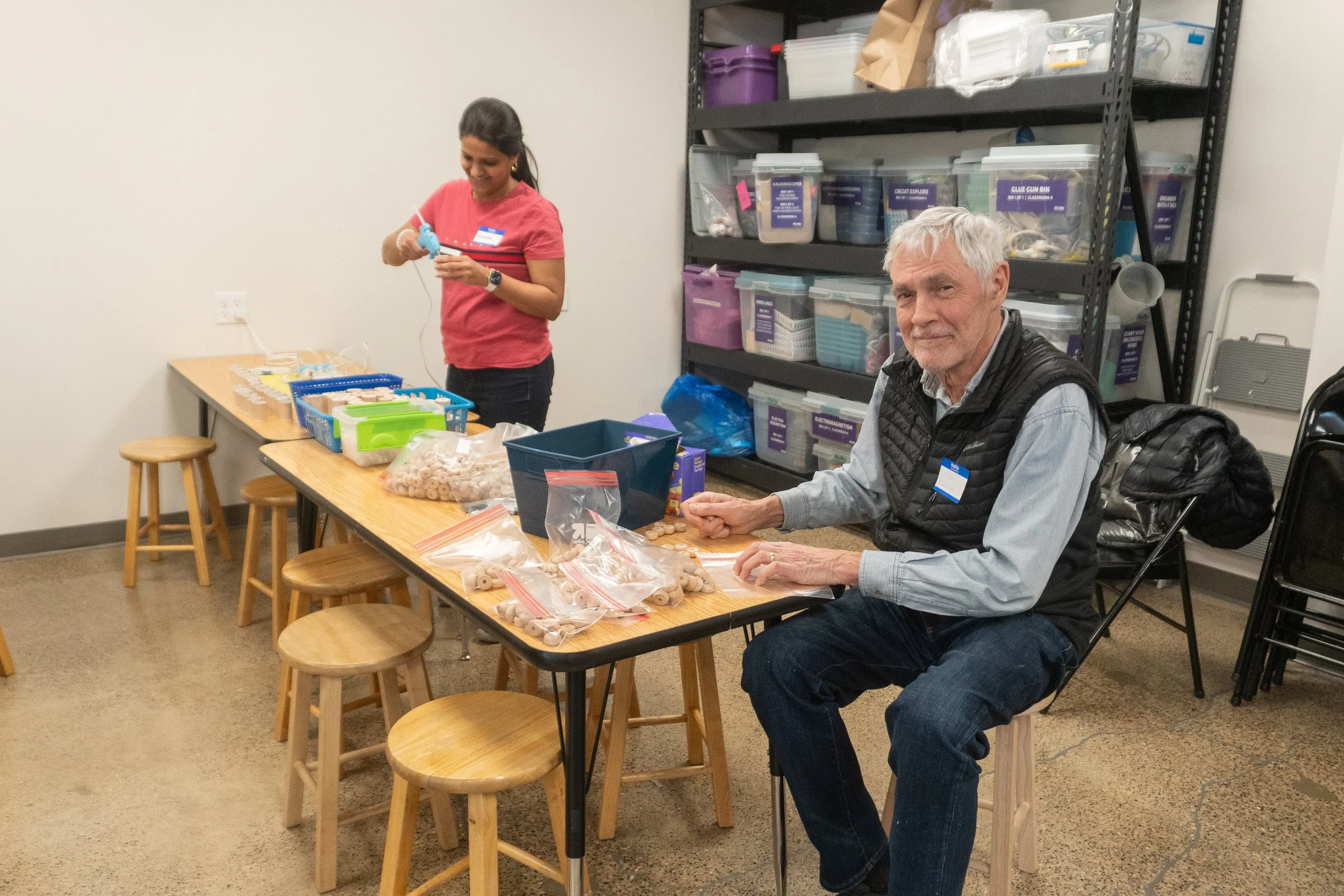 A woman and an elderly man are sitting at a wooden table, working on assembling or organizing small items in plastic bags. The woman is smiling and holding a small item, while the man is looking at the camera with a slight smile. Behind them is a metal shelving unit filled with storage boxes and supplies, indicating they are in a storage room or workshop setting.