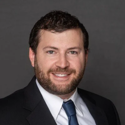 Portrait of a man in a business suit, smiling against a gray background.