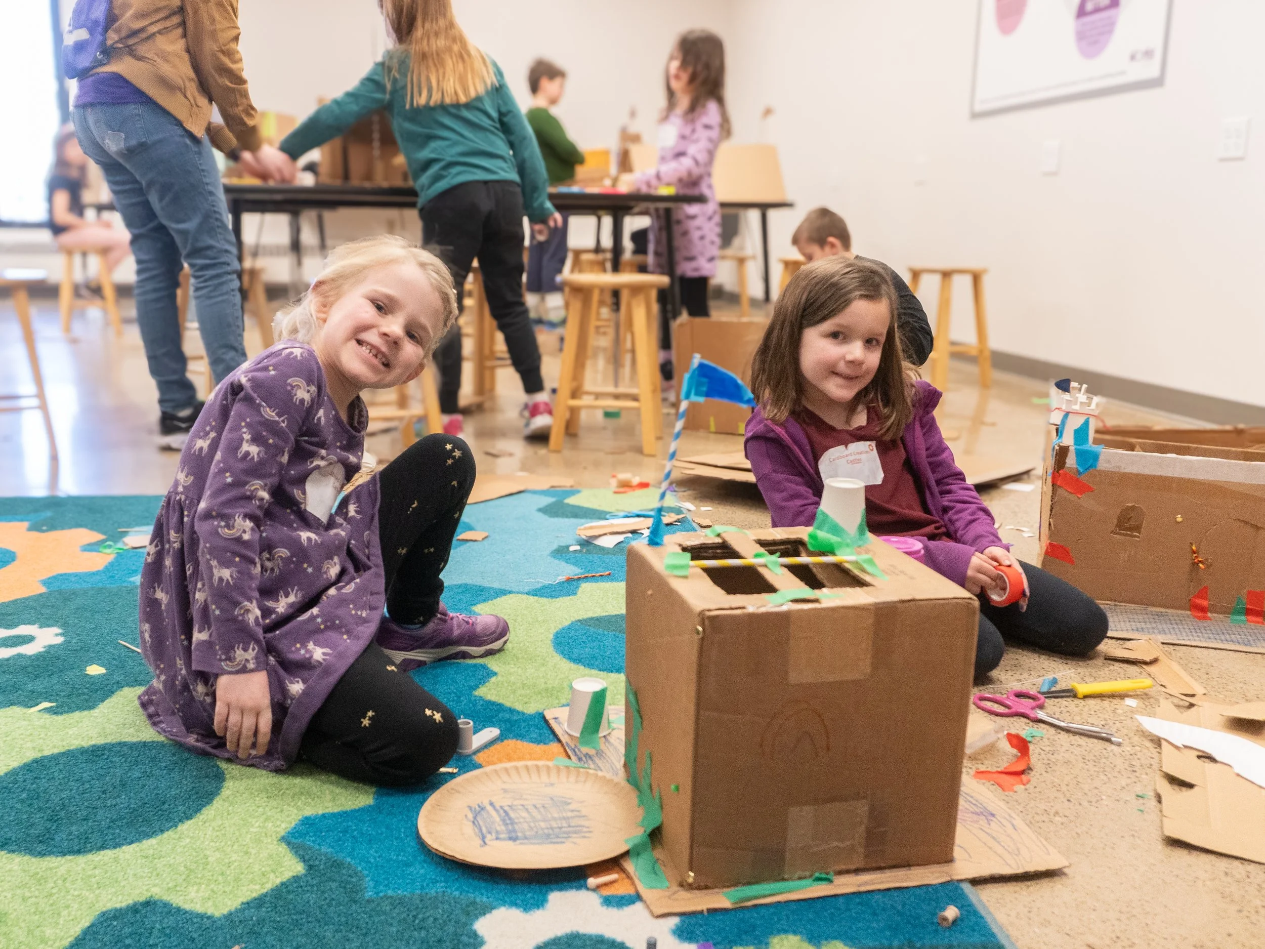 Two young girls sitting on a colorful rug, smiling at the camera, surrounded by cardboard crafts and art supplies, with other children and adults in the background participating in a craft activity.