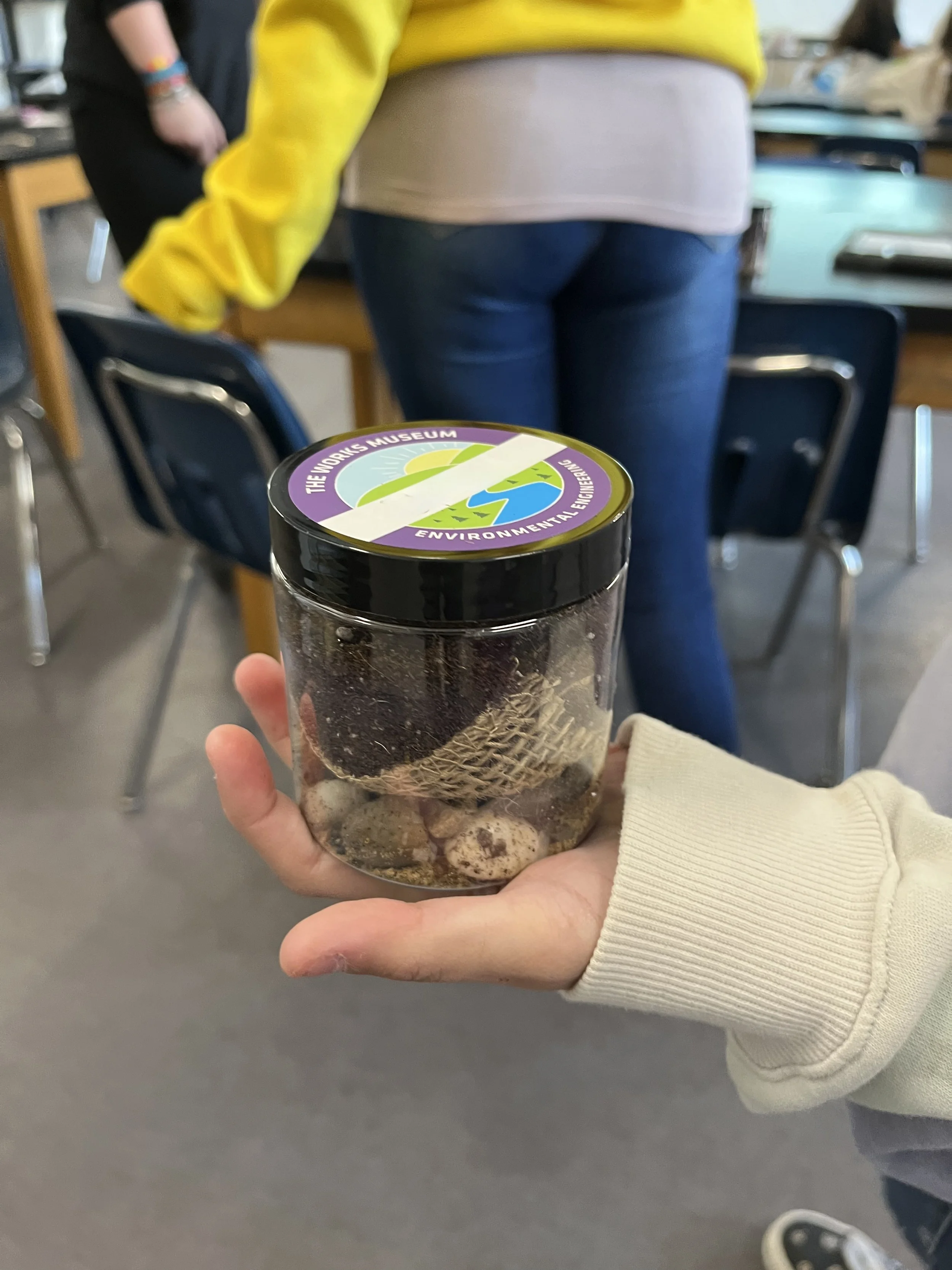 A person holding a jar with a label from The World Museum that reads "Environmental Engineering." The jar contains dirt, soil, or organic matter. In the background, there are chairs and a person wearing a yellow jacket.