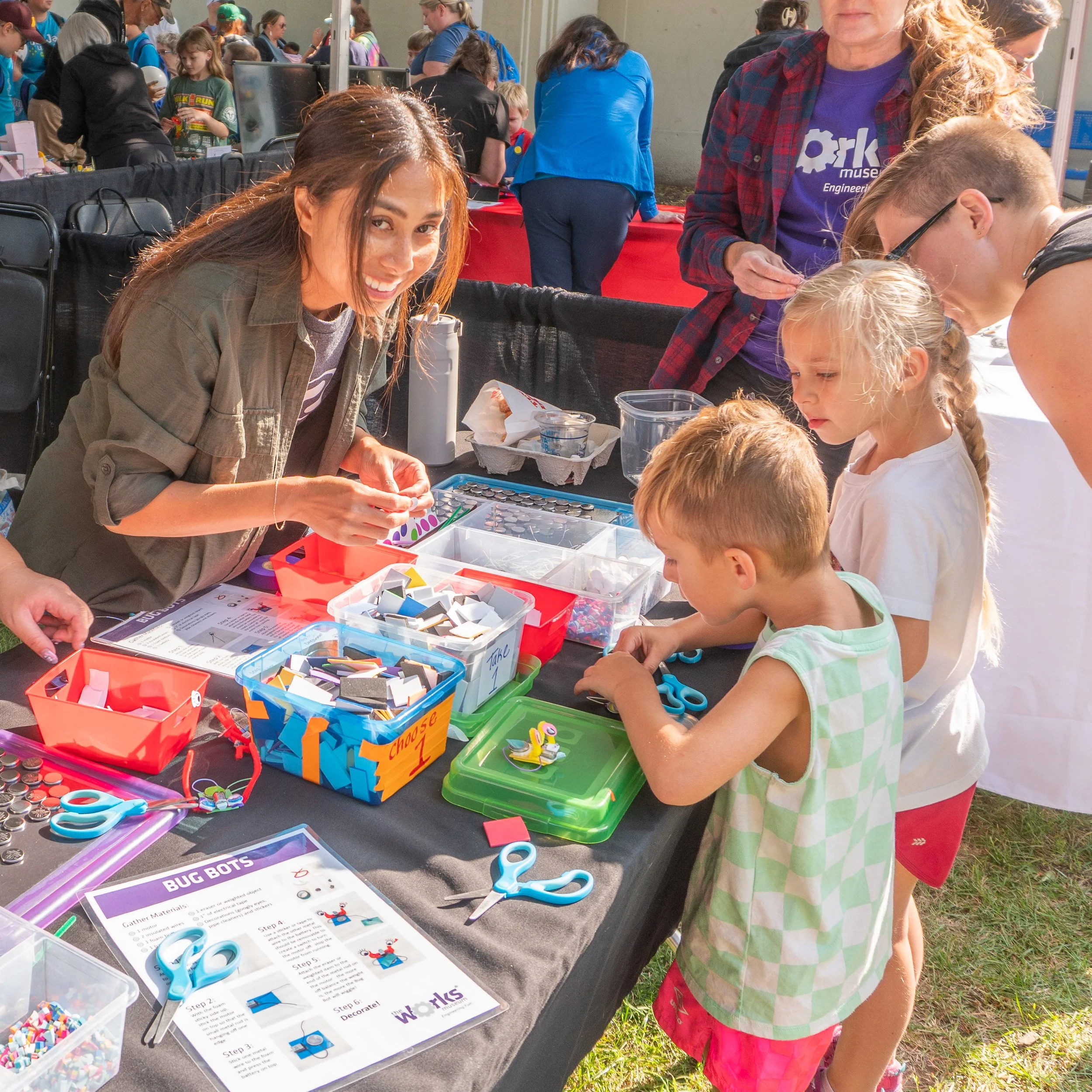Children and adults gathered around a table at a science or craft event outdoors, engaging with STEM activities involving colorful materials, scissors, and instruction sheets.