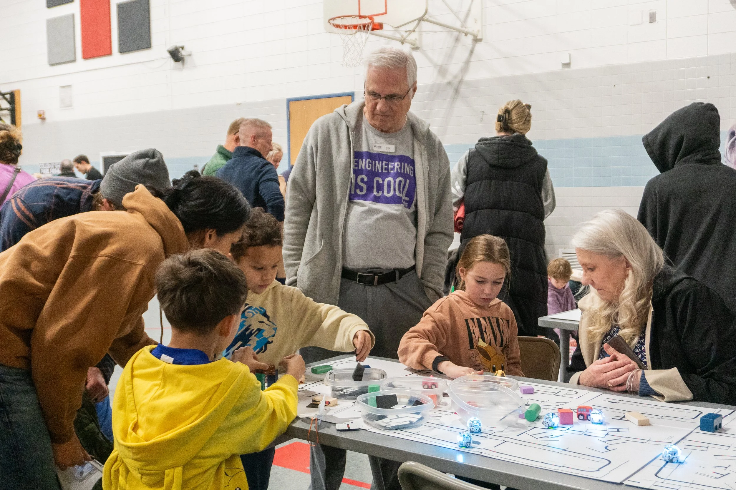 Group of children and adults gathered around a table in a gymnasium, engaged in a robotics activity with small robots and electronic components.