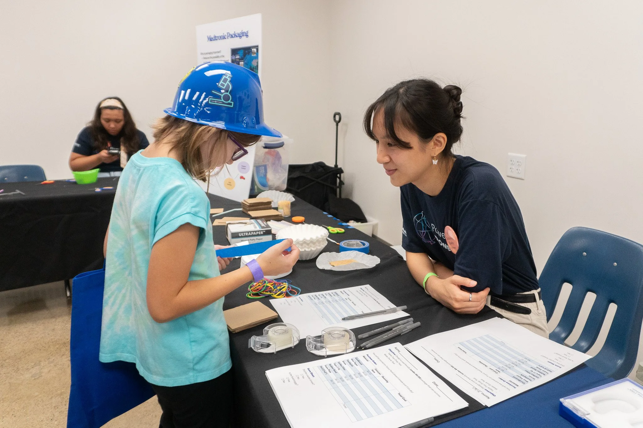 A young girl wearing safety goggles and a blue hard hat working on a project at a table, talking to an adult woman in a black T-shirt. They are in an indoor room with a black table covered in various materials, papers, and tools, and a poster on the wall behind them.
