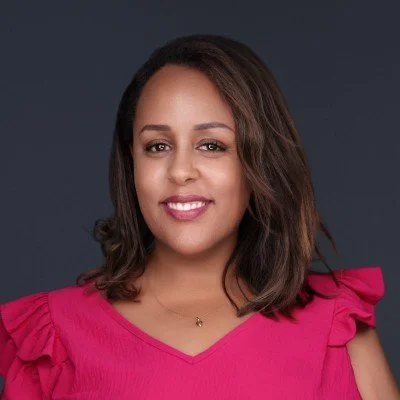 A woman with shoulder-length brown hair, wearing a pink top with ruffled sleeves, smiling against a dark gray background.