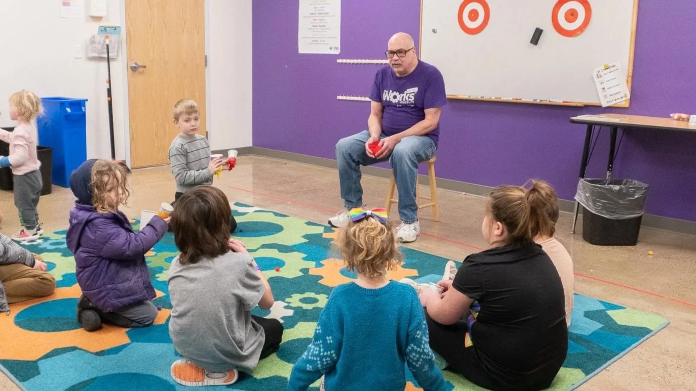 A classroom scene with a man sitting on a stool at the front, surrounded by children sitting or standing on a colorful rug. The man is holding a red object, possibly a small ball or toy, and the classroom has purple and beige walls with a whiteboard and targets in the background.
