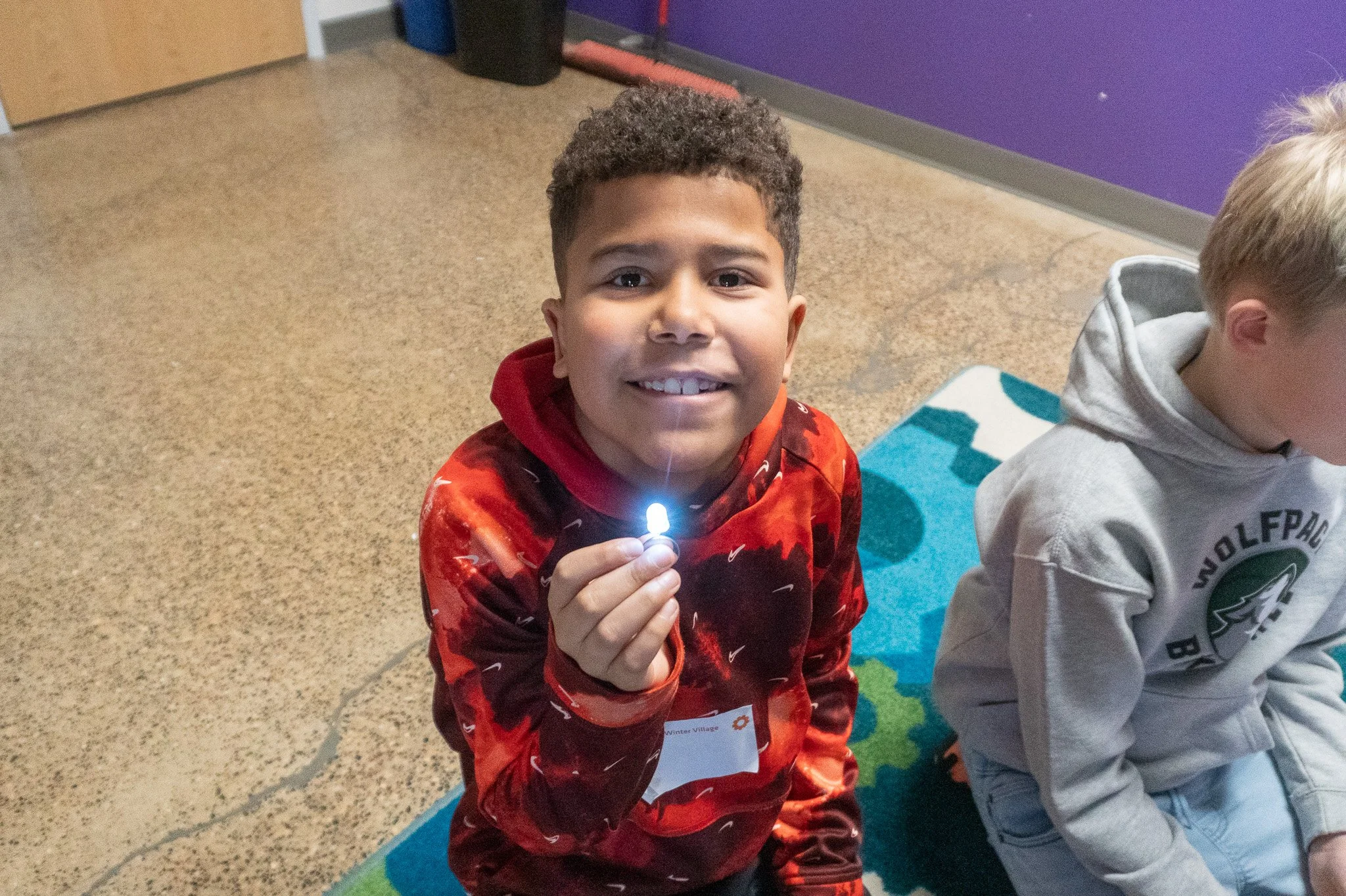A young boy with short curly hair smiling and holding a small lighted object, wearing a red and black patterned hoodie, sitting on a colorful rug inside an indoor space with a purple wall and a door in the background.