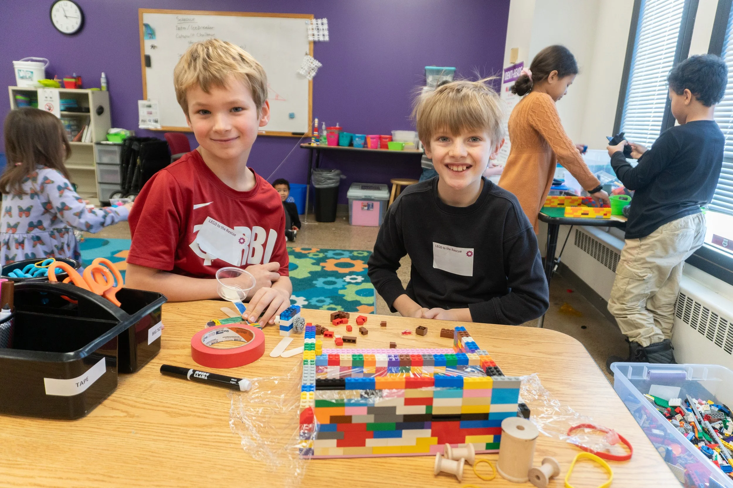 Two boys sitting at a table with a colorful Digital LEGO mosaic project, in a classroom with children in the background doing activities.