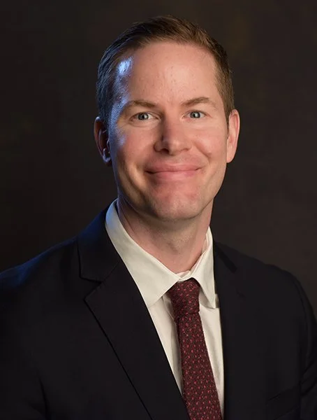 Portrait of a man in a suit with a white shirt and a red tie, smiling against a dark background.