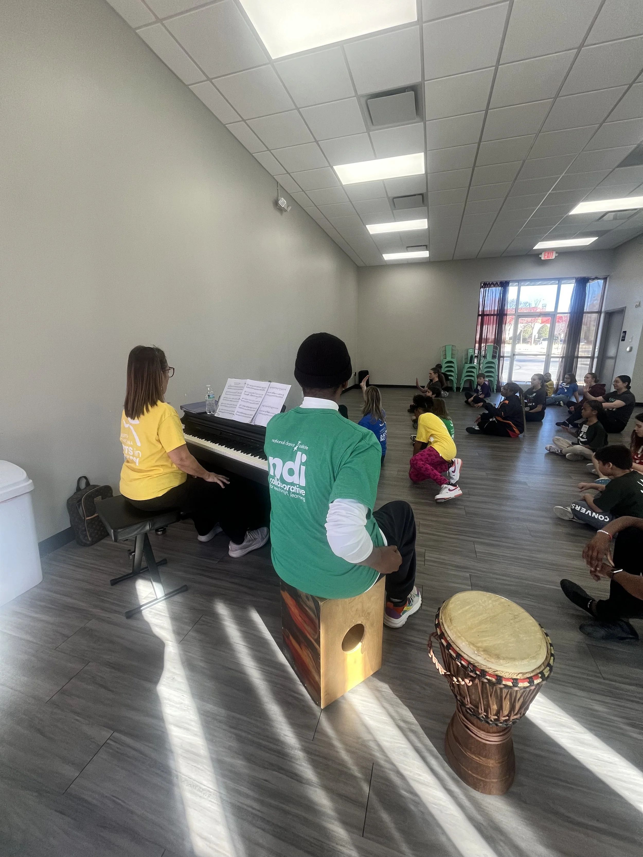 A group of children and adults seated on the floor in a classroom or community center, with a person playing a keyboard and another person playing a drum, leading a musical or movement activity.