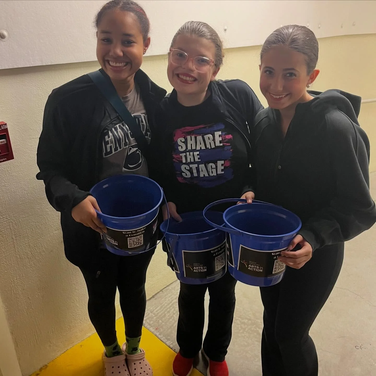 Three young women smiling and holding blue buckets, standing in a hallway with a yellow floor and a light-colored wall.