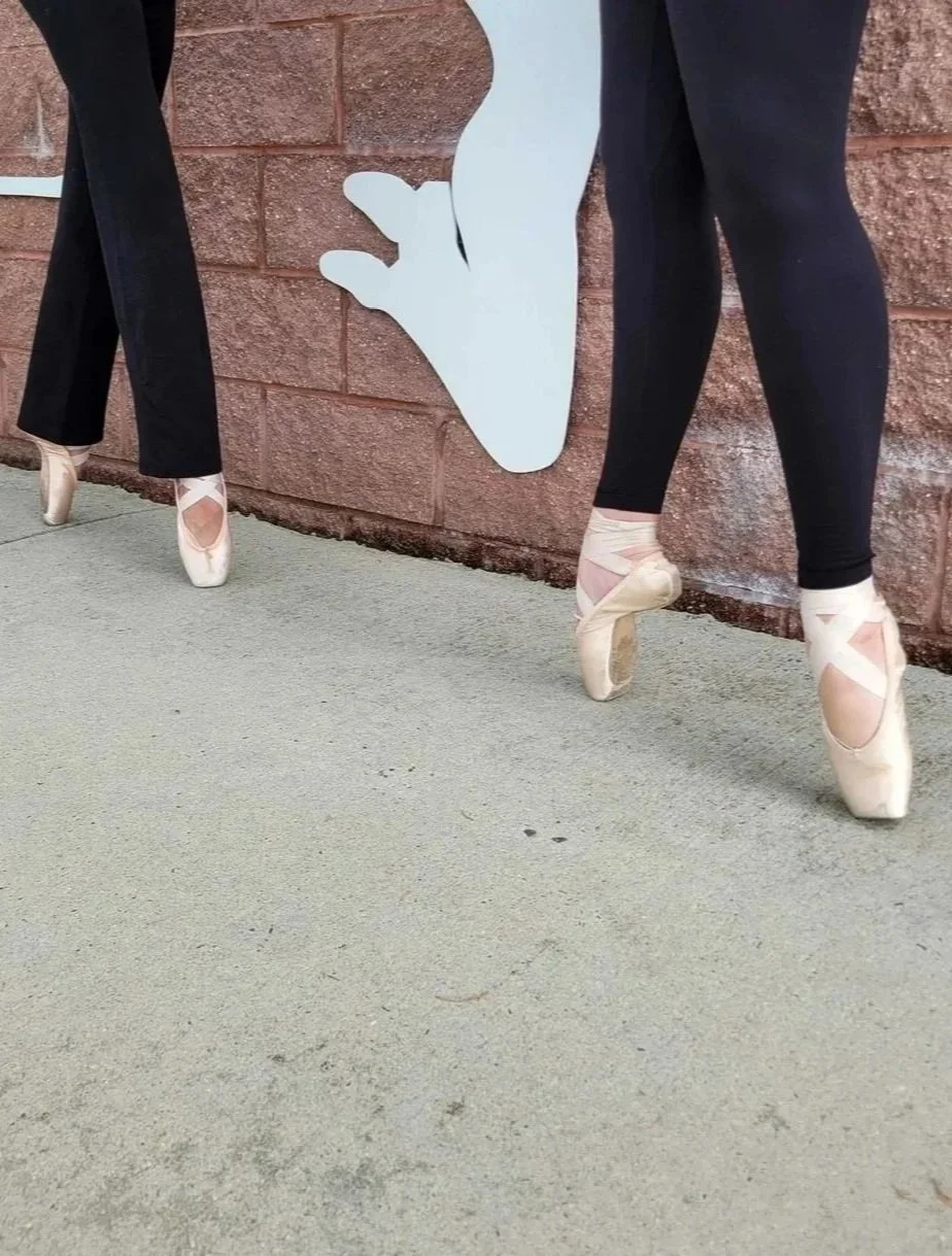 Two ballet dancers wearing pointe shoes with pink ribbons, standing on pointe, outside against a brick wall with a white silhouette of a hand on the wall.