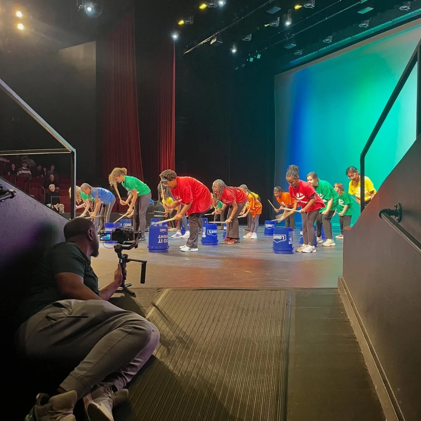 Children perform on stage by hitting drum buckets, dressed in colorful shirts, while a cameraman films from the side.