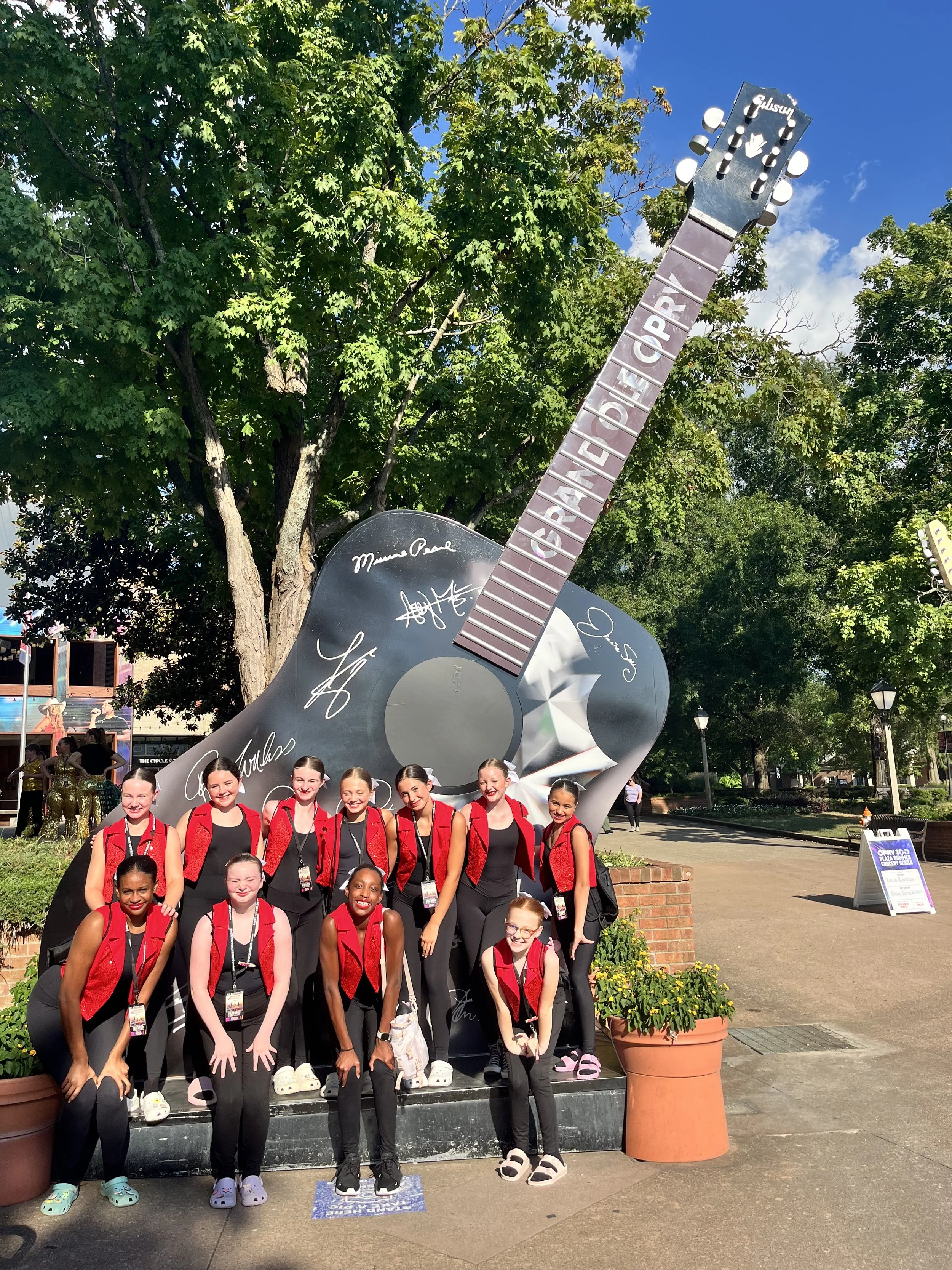 A group of young girls dressed in black with red vests, posing in front of a large guitar-shaped display that has signatures on it, outdoors on a sunny day with green trees and a blue sky in the background.