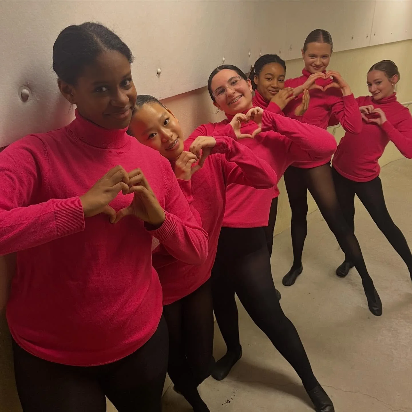 Group of six young women in matching red sweaters and black tights making heart shapes with their hands, standing against a wall in an indoor setting.