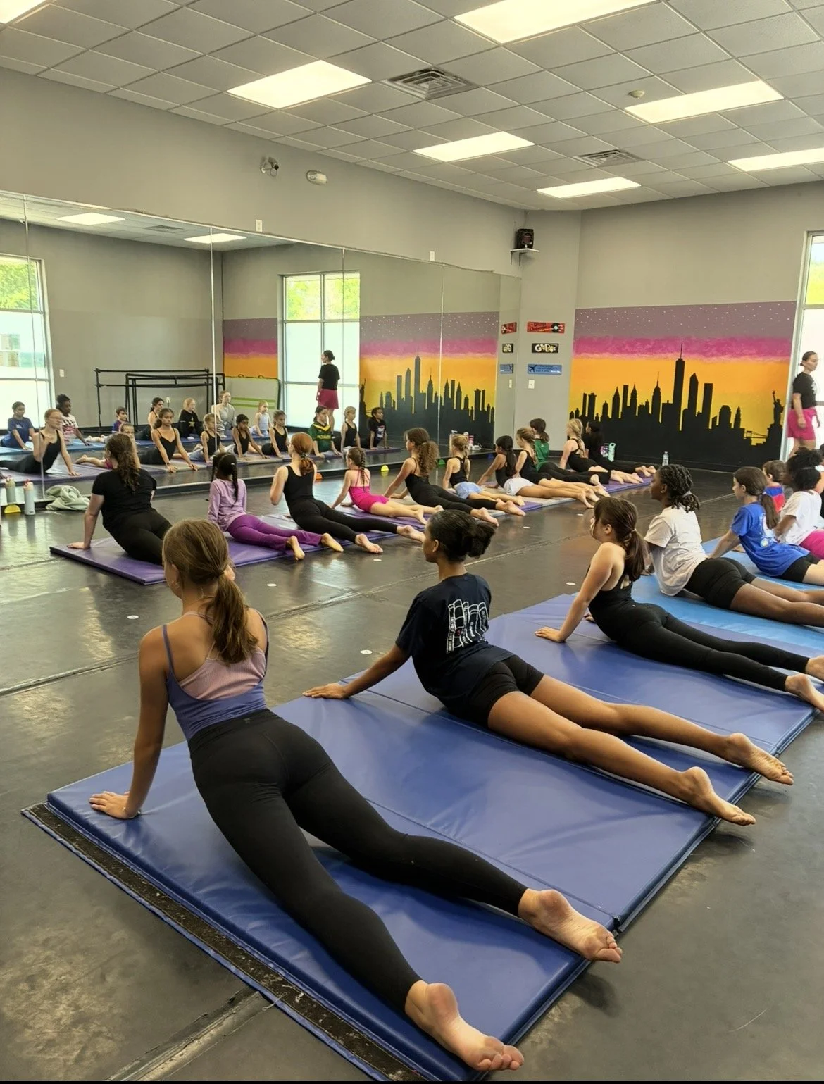 Young girls and women participating in a gymnastics class, performing stretches on mats in a dance studio with a city skyline mural.
