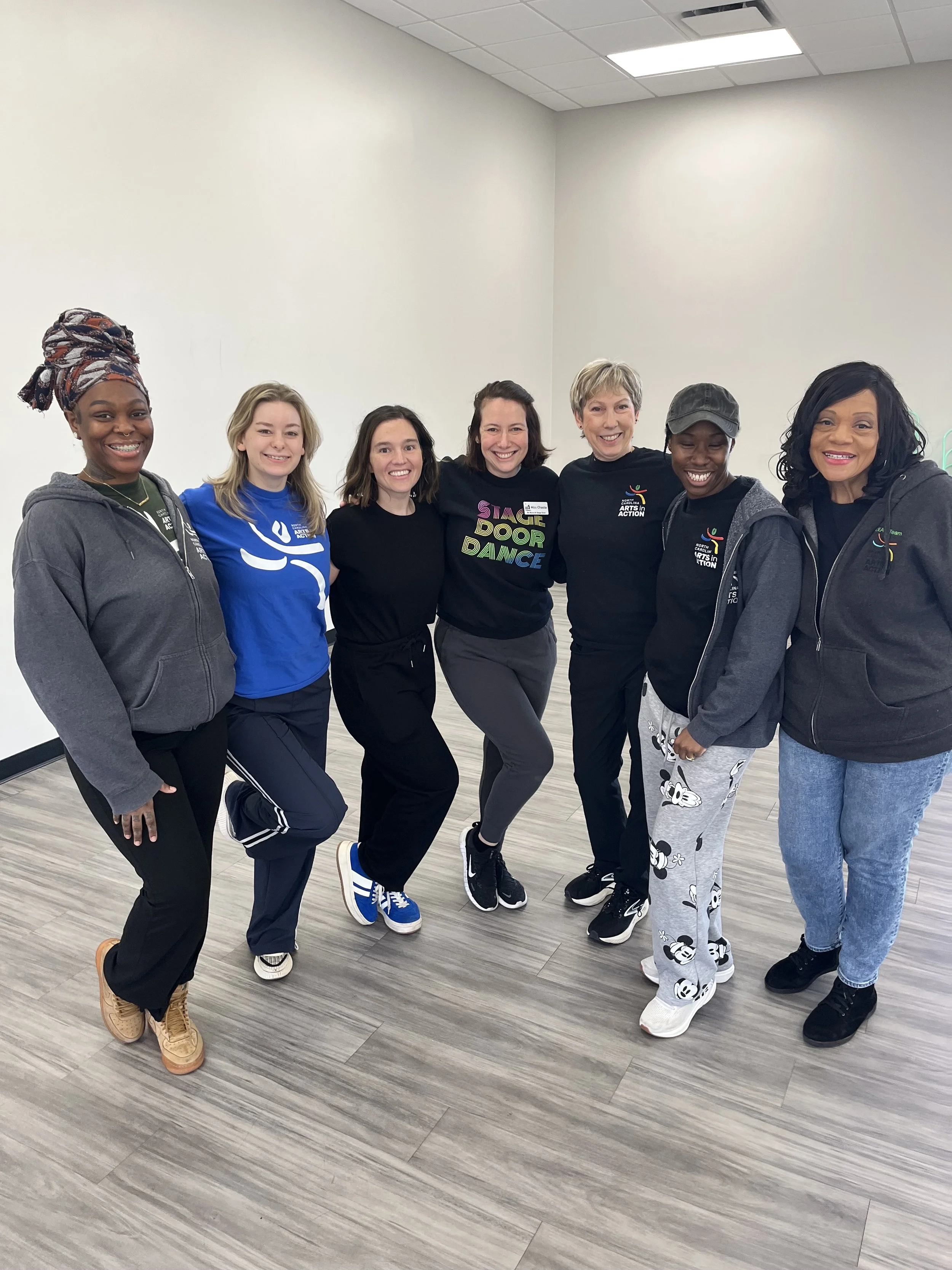 Group of seven women standing together indoors, smiling, wearing casual and athletic clothing, in a plain room with a light-colored wall and wood flooring.