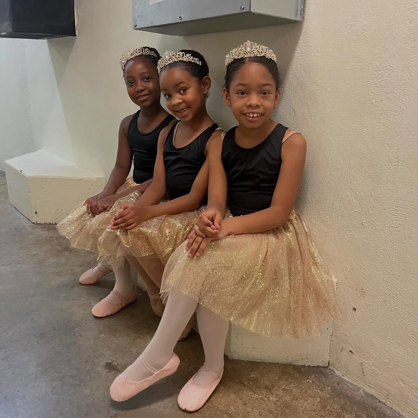 Three young girls sitting on a bench, wearing ballet costumes with black tops, gold skirts, pink tights, ballet shoes, and tiaras, smiling at the camera.