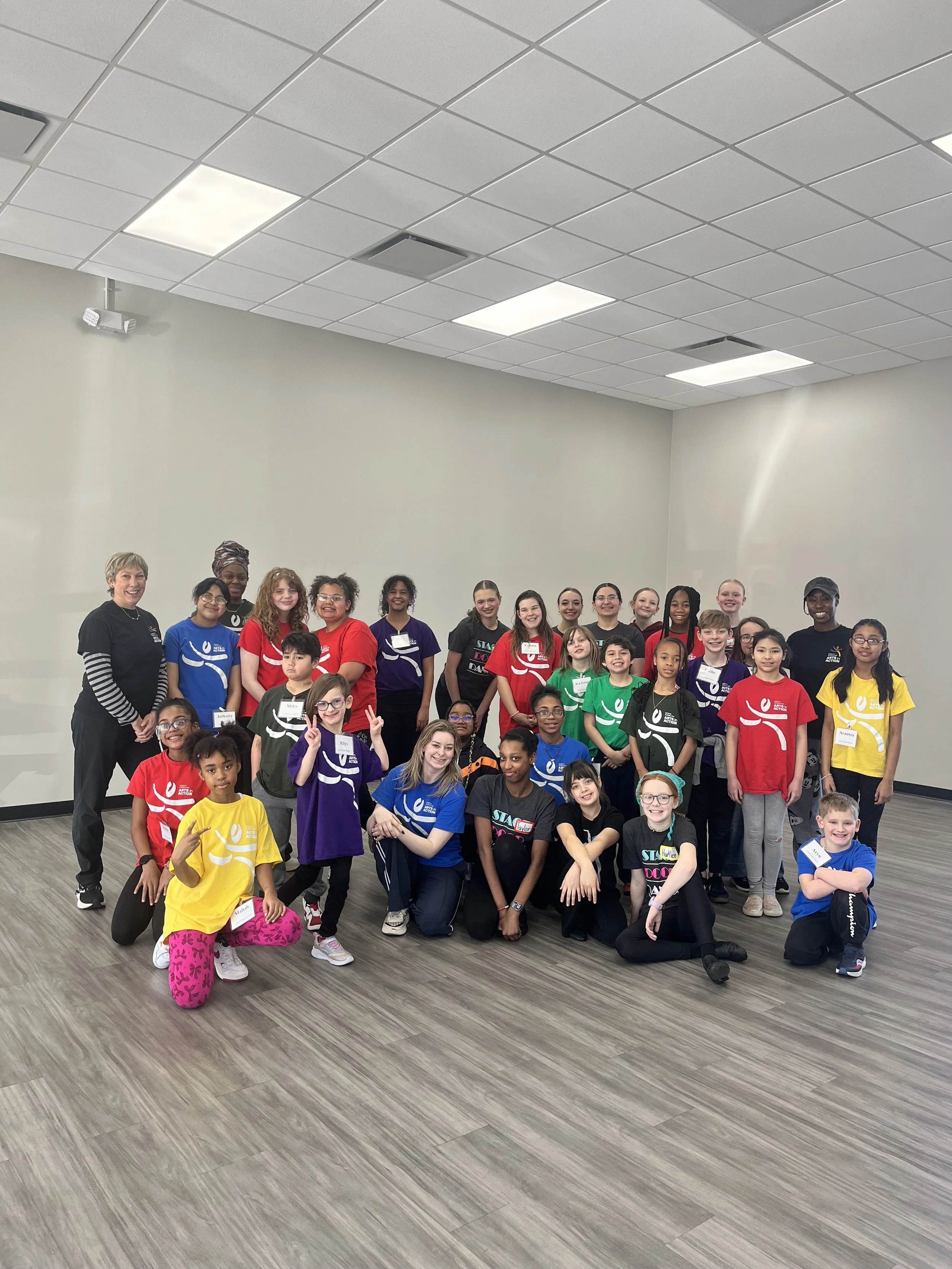 A group of children and adults posing for a photo in a room with a gray floor and plain white walls, some wearing colorful T-shirts with a logo.