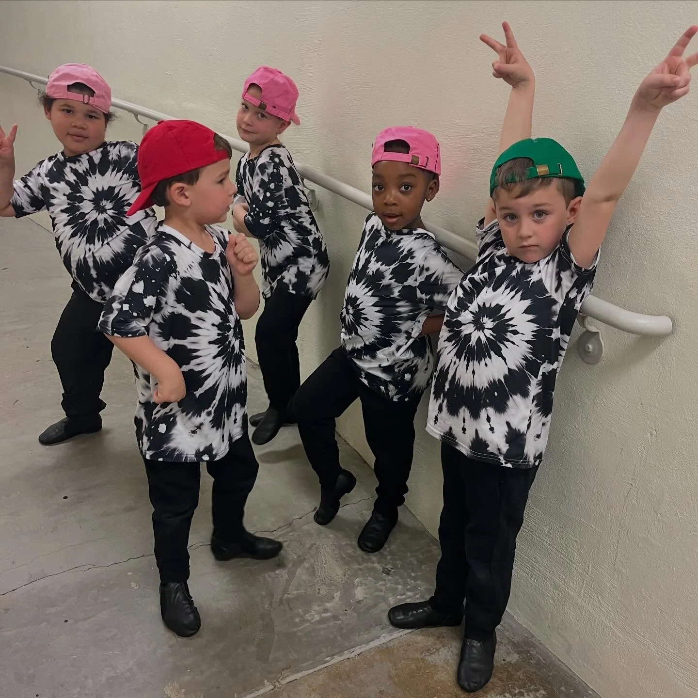 Group of five children wearing matching black and white tie-dye shirts, with colorful caps, posing against a wall and handrail.