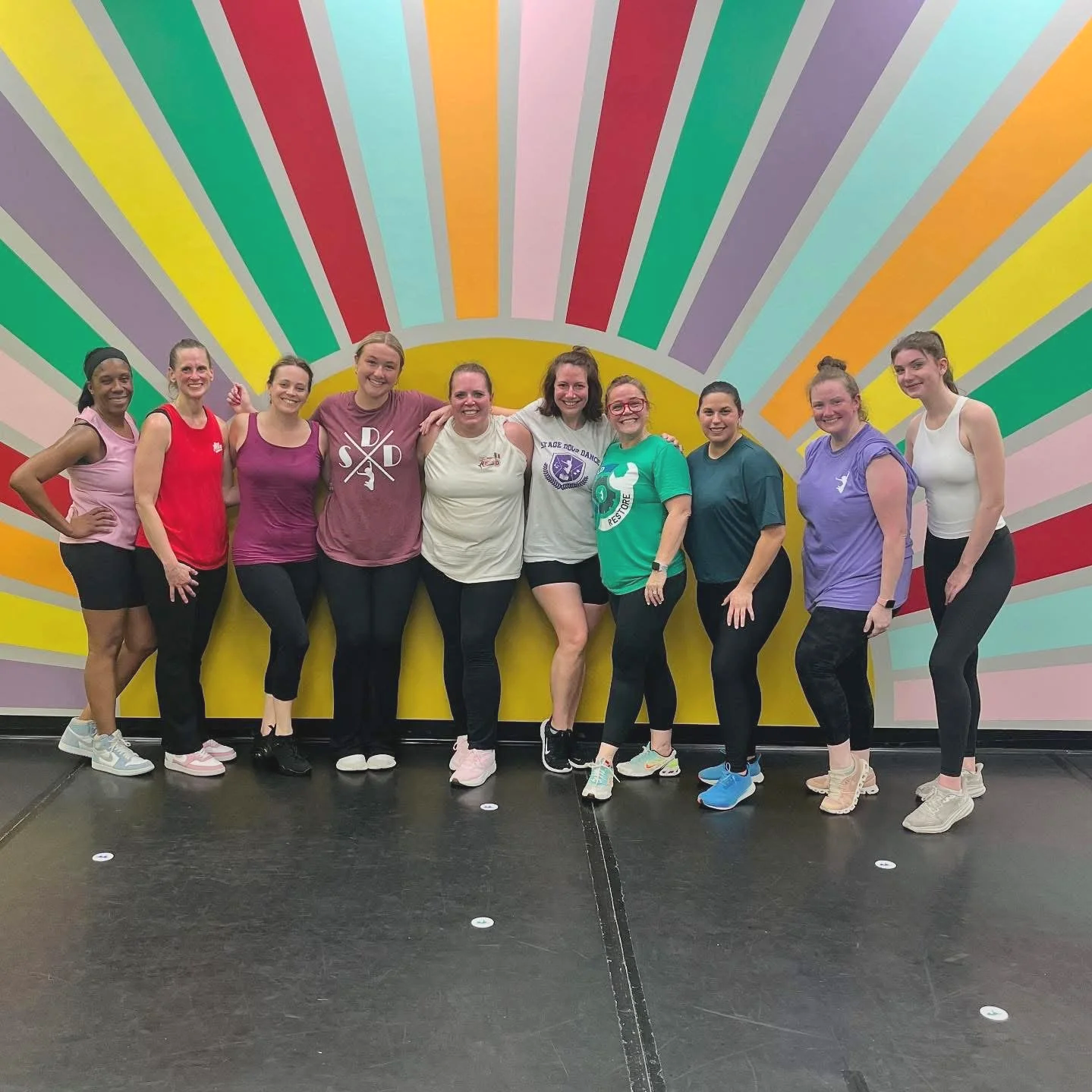 A group of ten women standing together in a dance studio with a colorful striped mural background, smiling and posing for a photo.