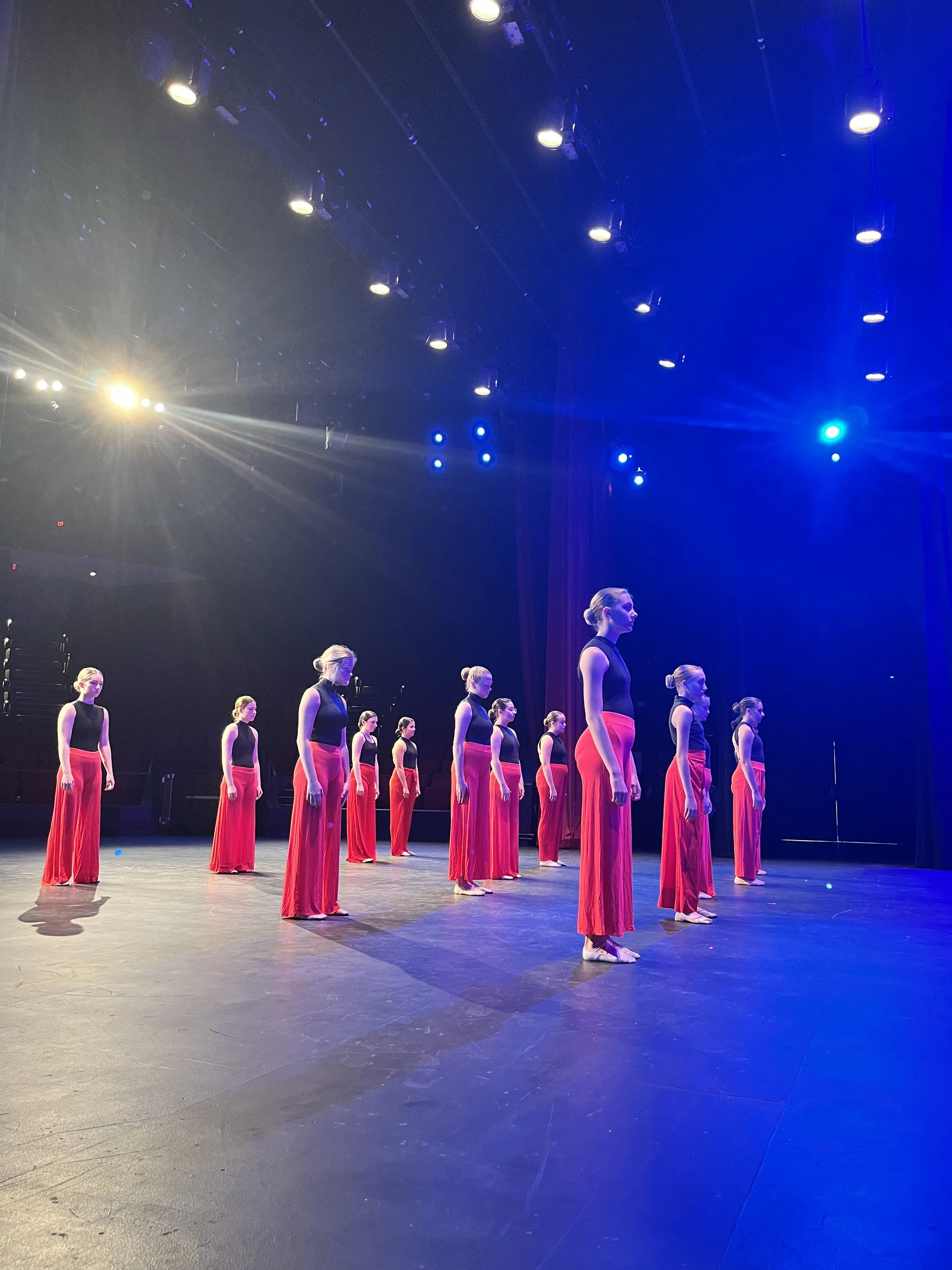 Dance performers in black tops and red wide-leg pants standing in a formation on a stage, illuminated by stage lights with a dark background.