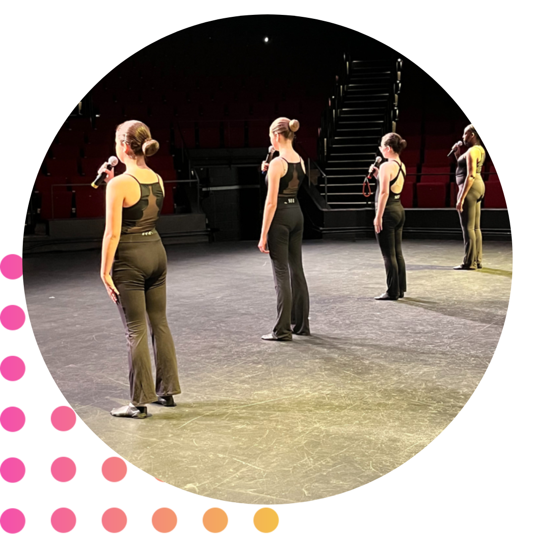 Five women on stage, standing in a straight line, each holding microphones, in a theater or performance space with dark background and seating.