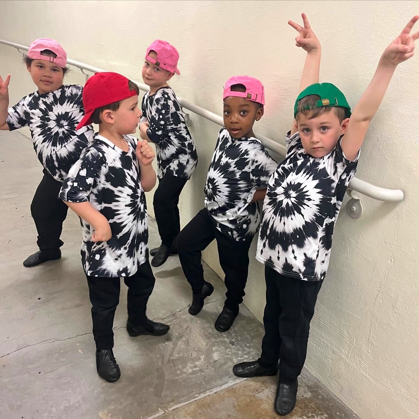 Six children wearing matching black and white tie-dye shirts, black pants, and black shoes. They are wearing colorful caps, including pink and green. The children are posing with peace signs and various expressions, standing close to a railing against a beige wall.