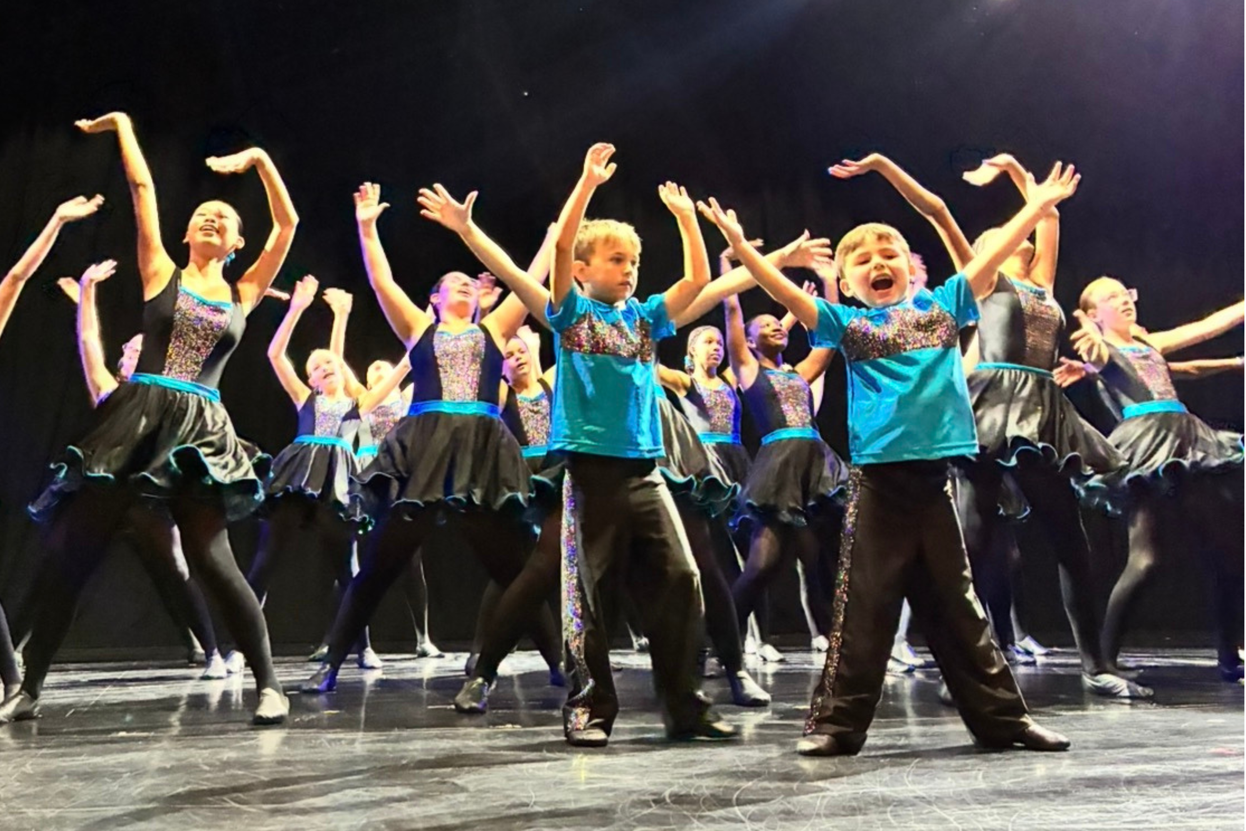 Children performing a dance on stage, wearing coordinated black and blue costumes with embellishments, with arms raised and smiling expressions.