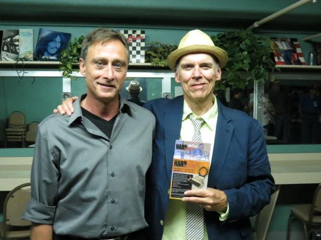 Gary Appleagate and John Hiatt are standing together in the green room after John's musical performance. John is wearing a light-colored hat, blazer, and tie, and holding a HarpLock box. Both men are smiling.