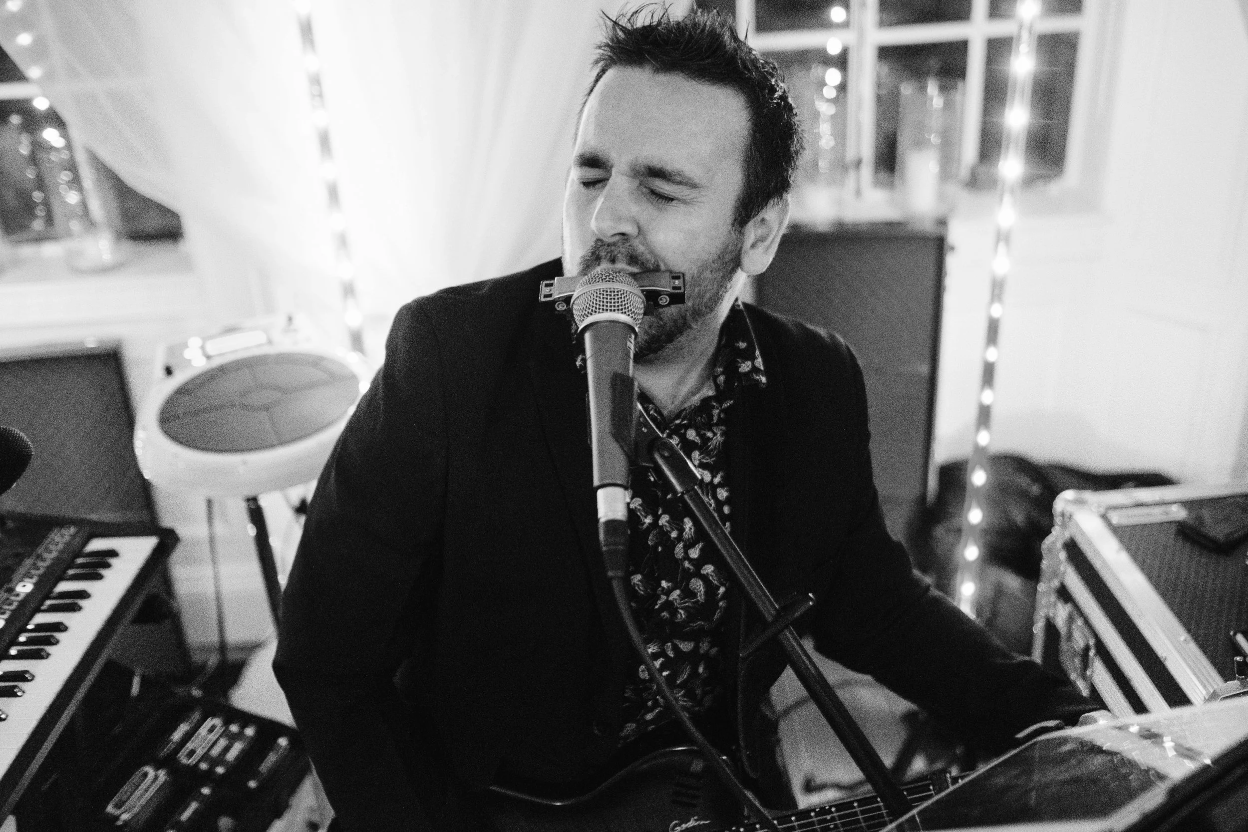 A Matt O'Neill playing harmonica into a microphone in a music studio, surrounded by musical instruments including keyboards and guitars, with string lights in the background. He is using a HarpLock to hold the harmonica in place in front of the mic.
