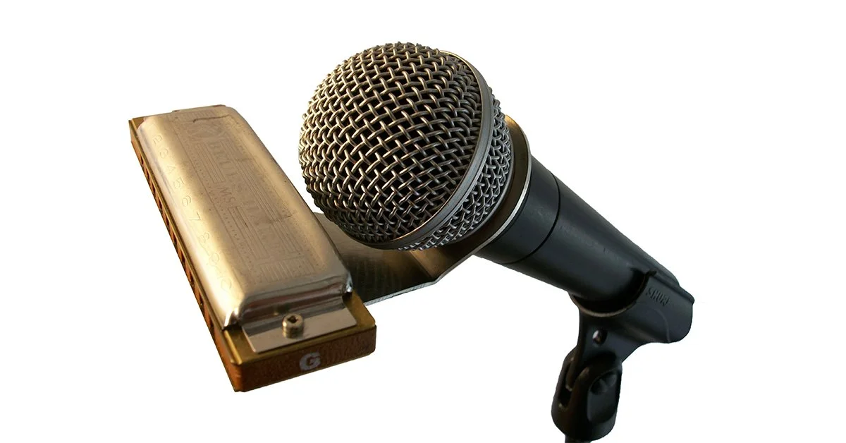 A microphone with a large circular metal grille, a black handle, and a mounted harmonica in a white background.