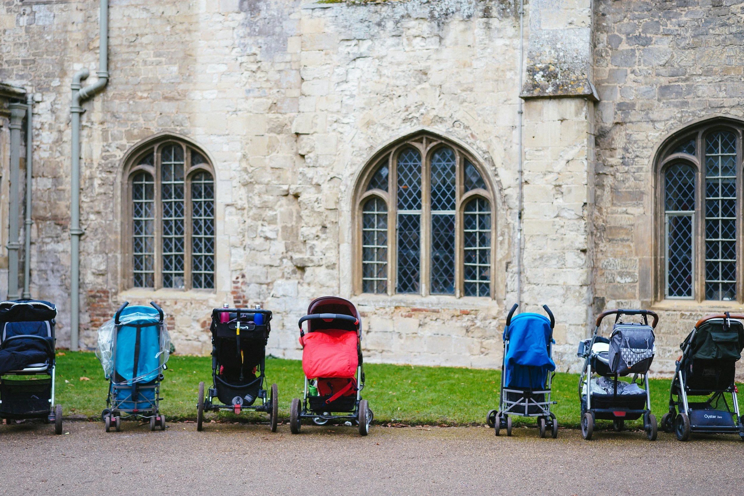 A range of strollers are lined up outside a building which could be an old church. You can see there is a wide variety of strollers.