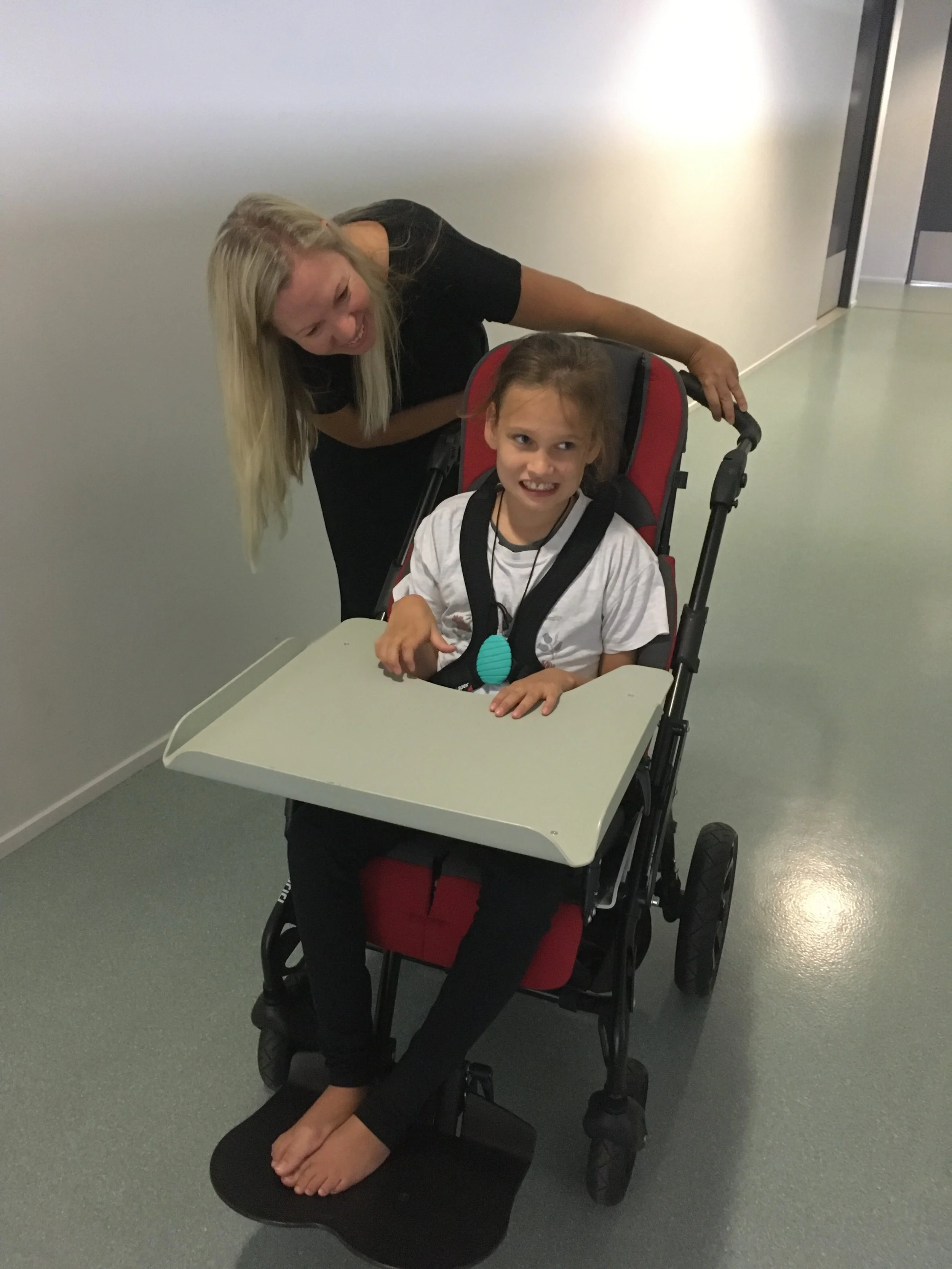A young girl in a wheelchair smiling with a caregiver leaning over her in a hallway during assistive technology trials.
