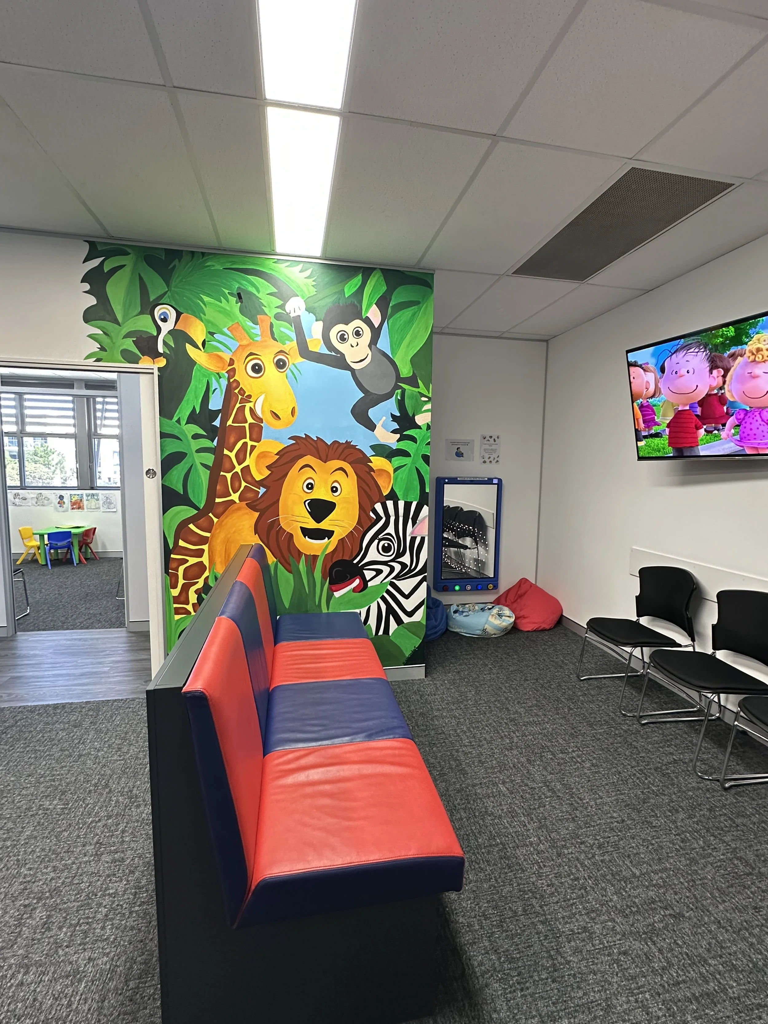 A jungle mural adorned the wall of a clinical waiting room. You can also see beanbags, a sensory mirror and TV playing cartoons. Through he door you can see into the OT office with children's table and chairs.