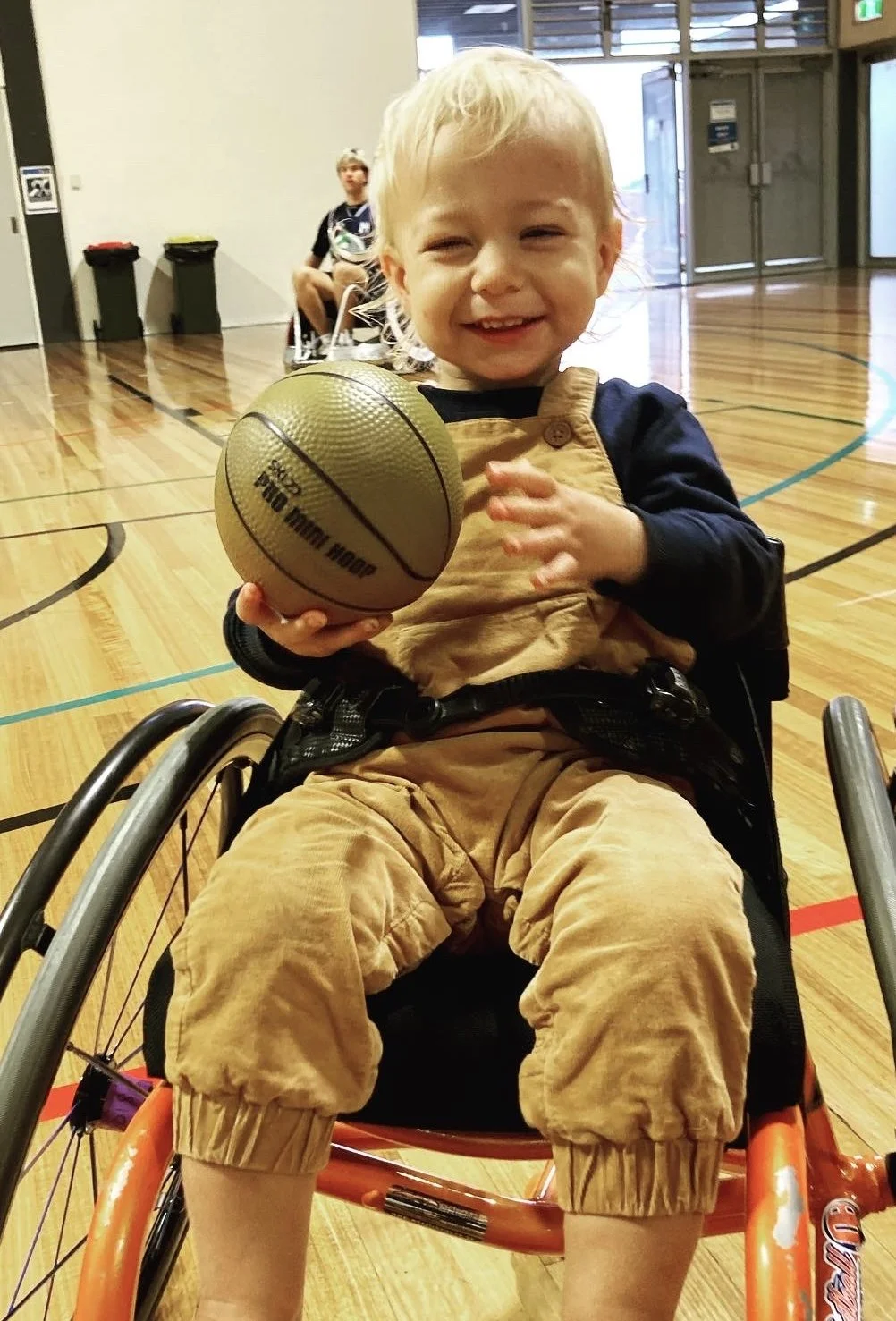 A cute blonde toddler is holding a miniature basketball and smiling at the camera. He is sitting in a basketball wheelchair.