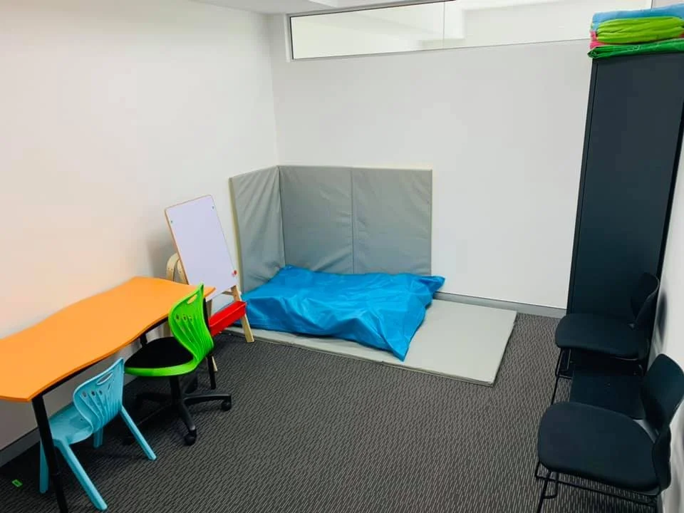 Sippy Downs Clinic - A small inviting room with a crash mat on the floor, green and blue chairs and table, a small whiteboard, and a closet full of games!