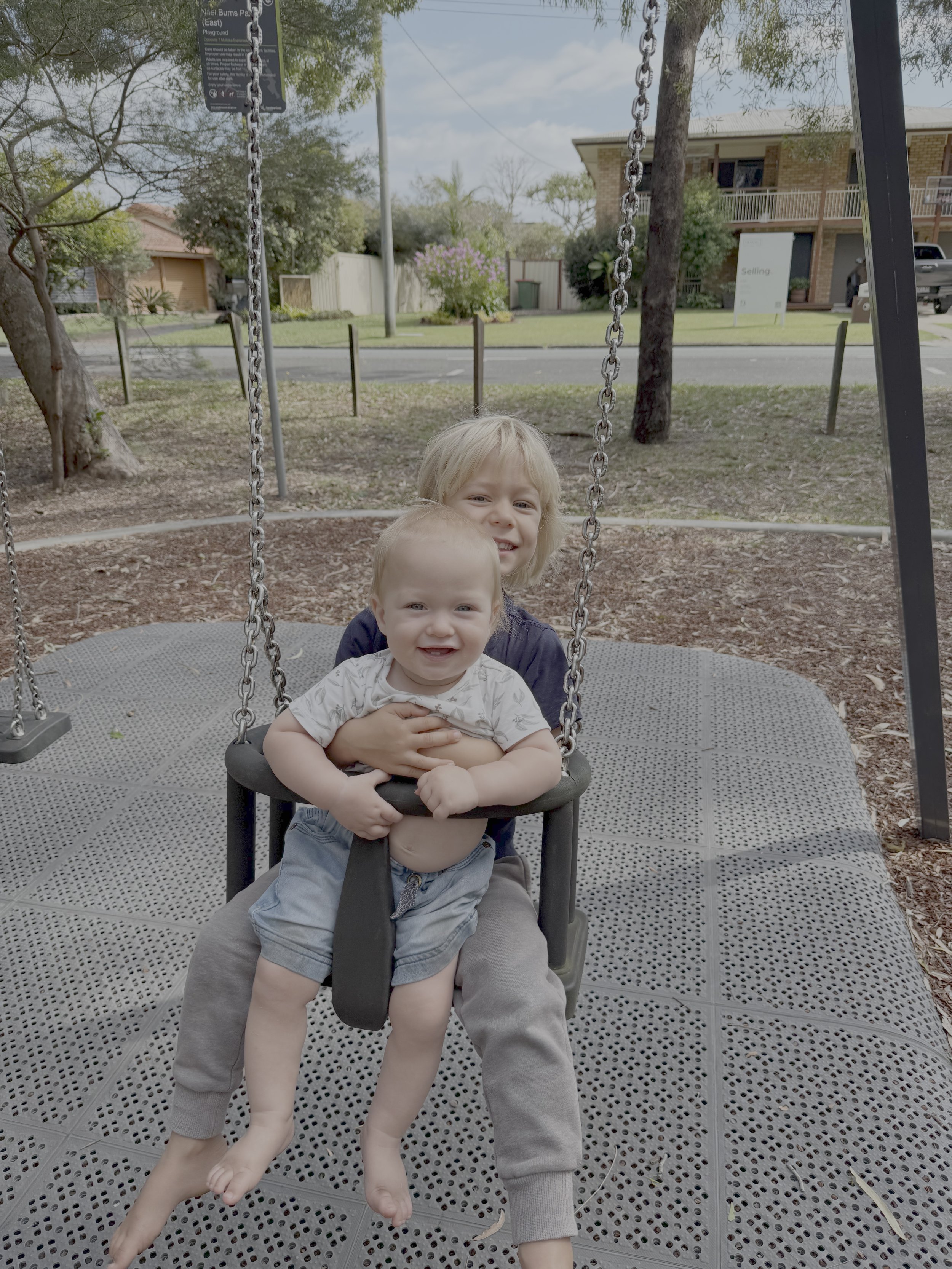 Two children, one older and one younger boy, sitting on a swing set at a park.
