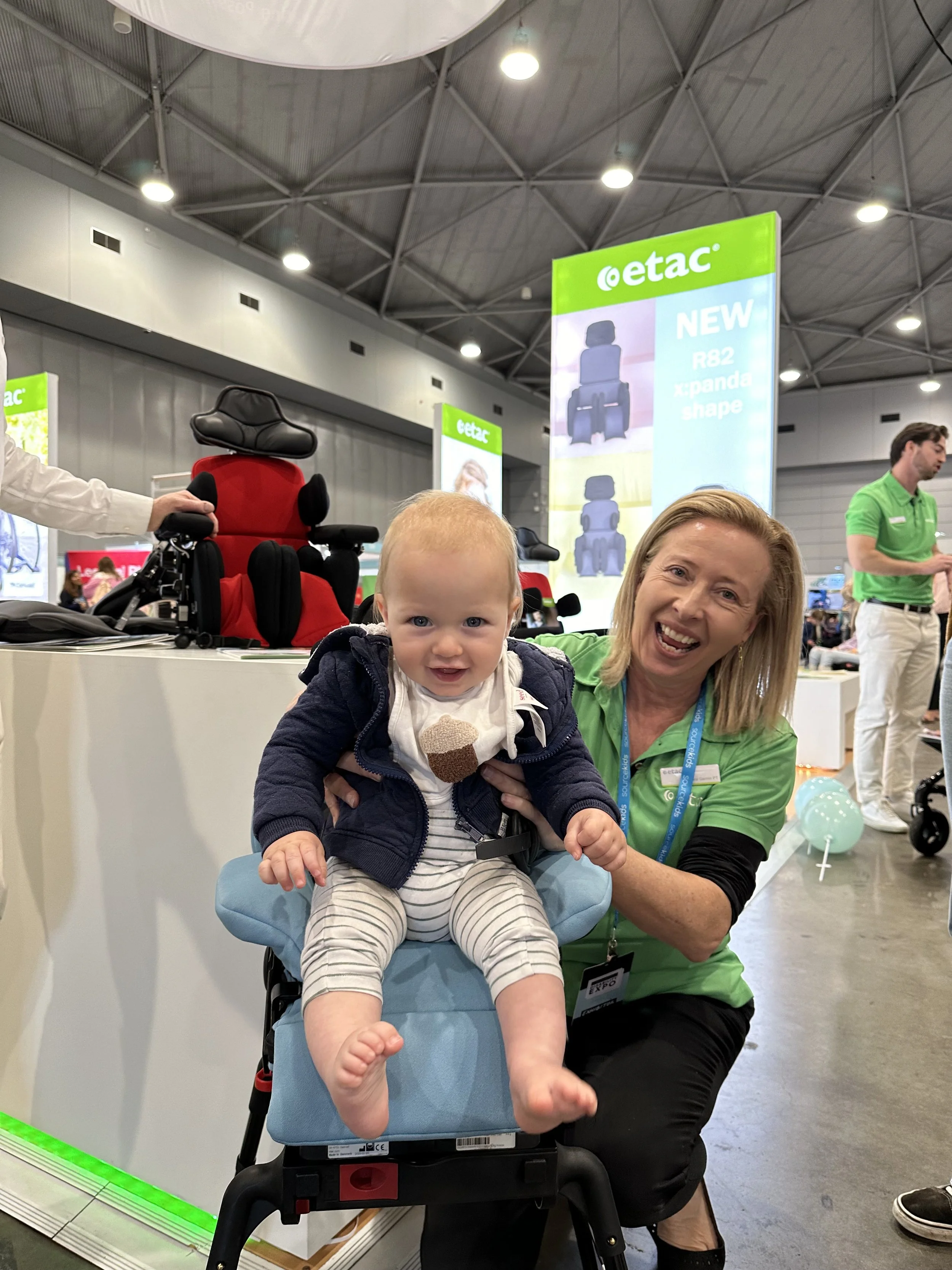 A woman with blonde hair smiling and holding a young baby in supportive seating at a trade show. The background features a children's car seat, along with other people and display booths.