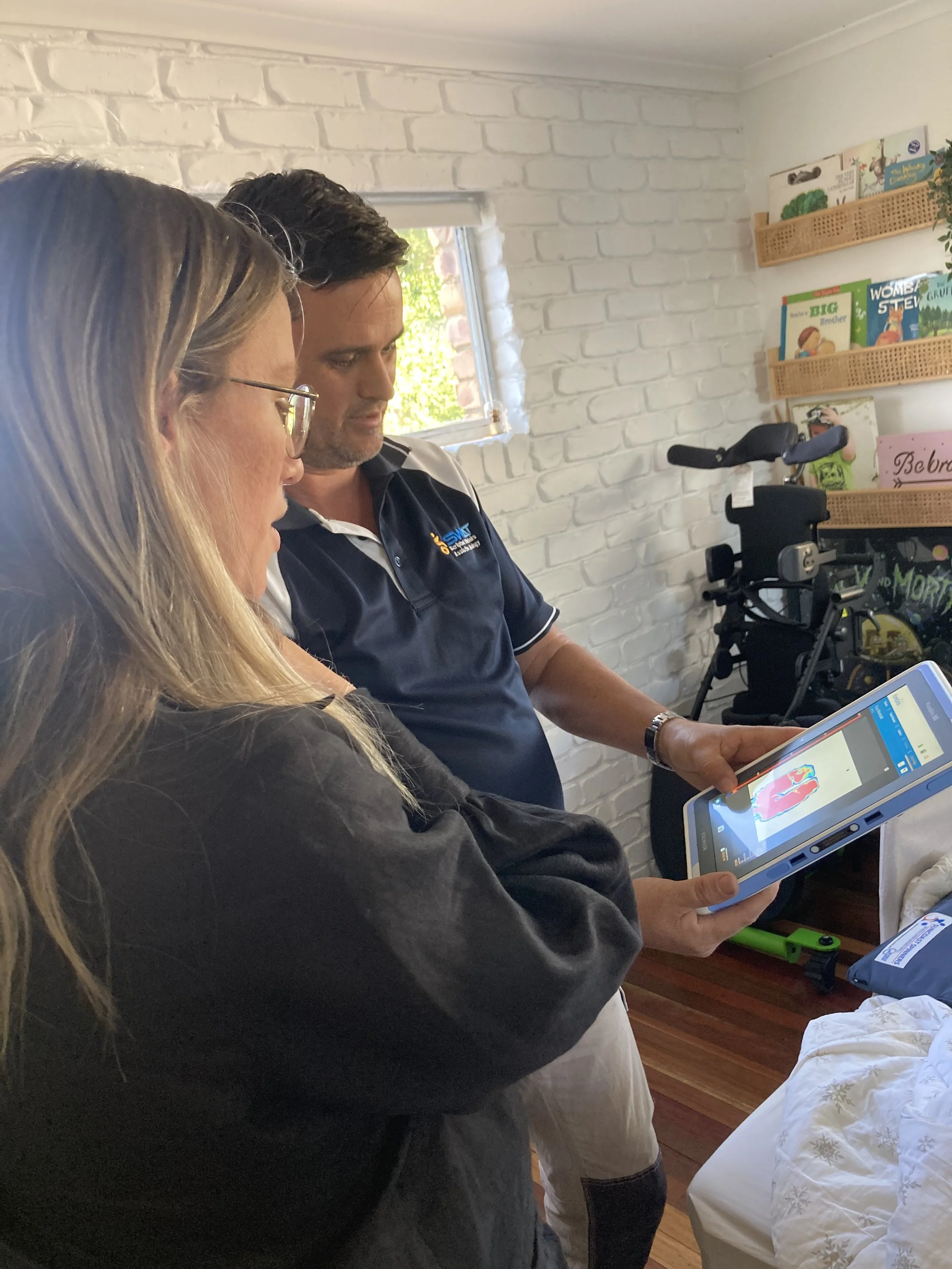 Two people, a woman with long blonde hair and glasses, and a man with dark hair, are looking at a tablet together in a room with white brick walls. The woman is holding the tablet, and they are focused on the screen, which displays a colorful image. Behind them are shelves with children's books, a walker, and a window showing greenery outside.