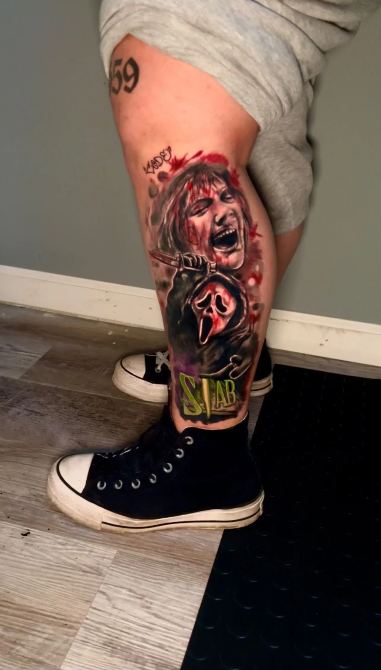 Tattoo on person's leg depicting a woman's face screaming with blood, holding a knife, and wearing a hooded figure with a masked face below. The tattoo also features the word 'STAB' at the bottom.