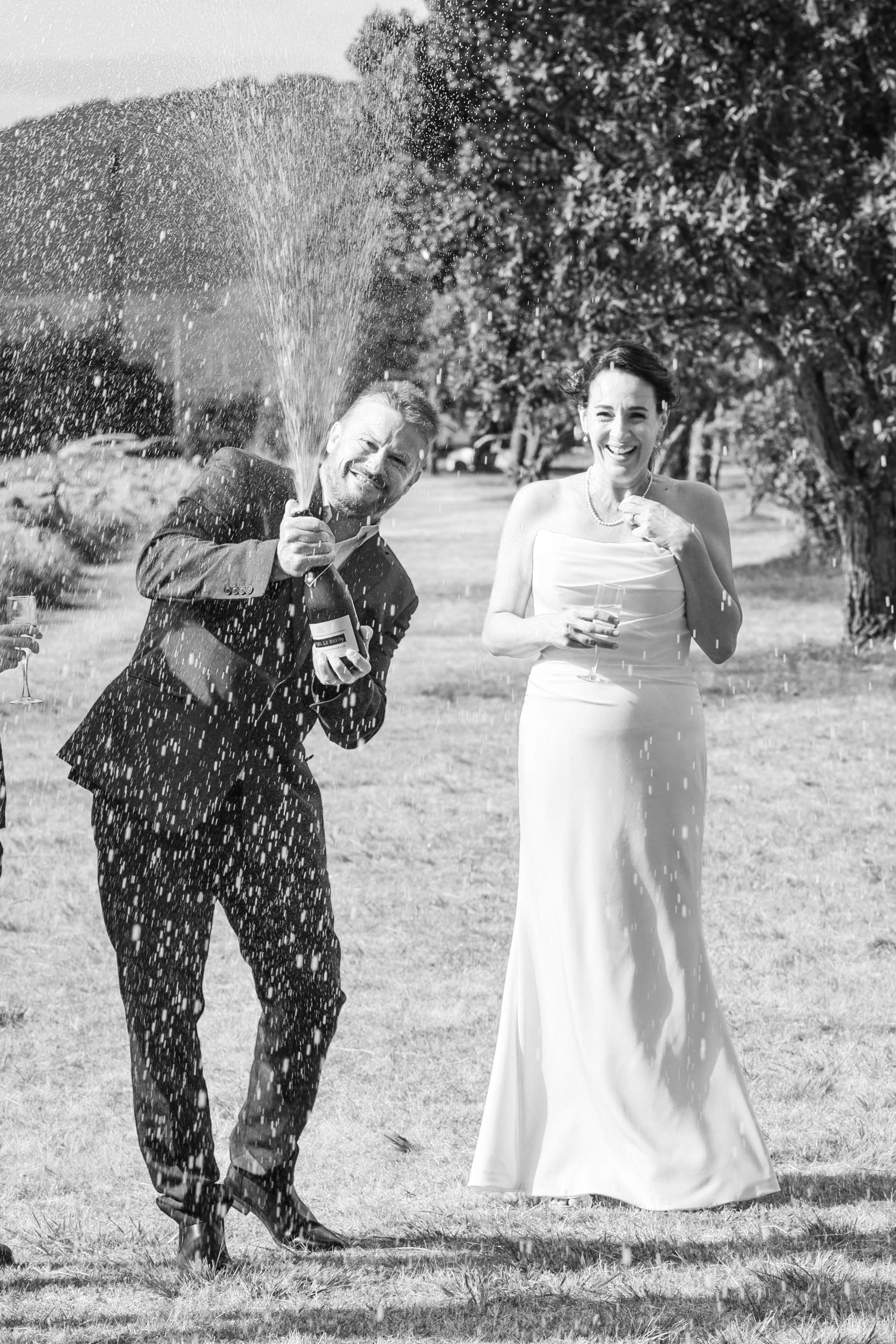 Black and white photo of a man and woman celebrating outdoors. The man is wearing a suit and popping a champagne bottle, while the woman is in a strapless dress holding a glass. They're surrounded by trees and grass.
