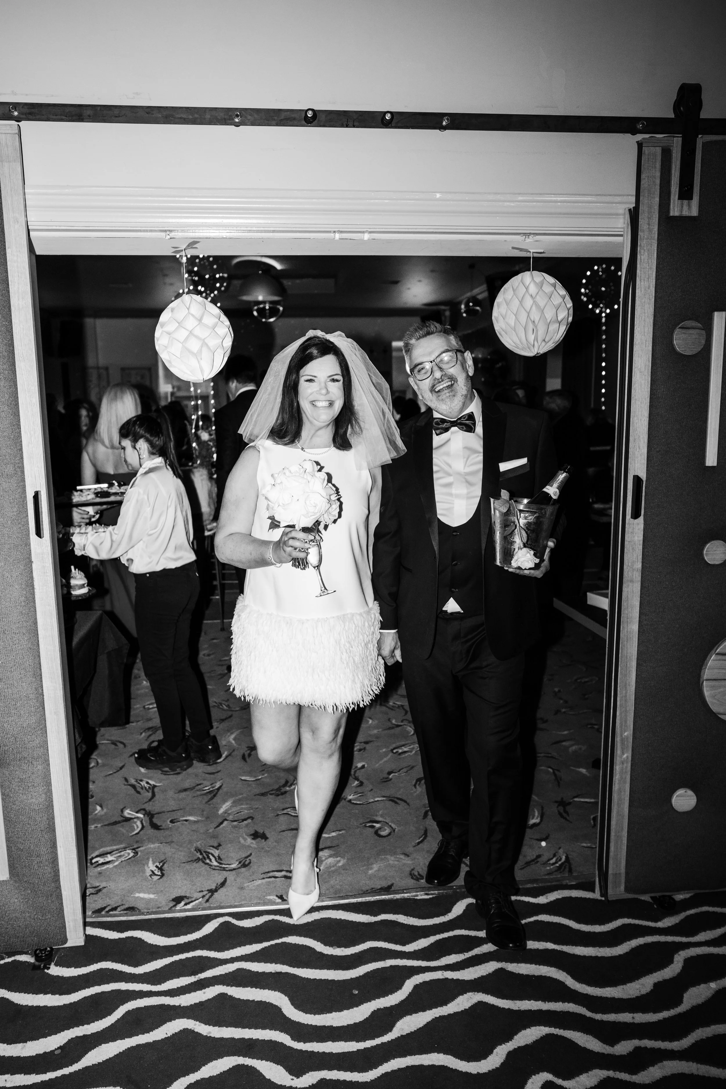 Black and white photo of a woman and man in wedding attire walking into a celebration, smiling and holding flowers and a gift. The background shows guests, hanging paper lanterns, and festive decor.